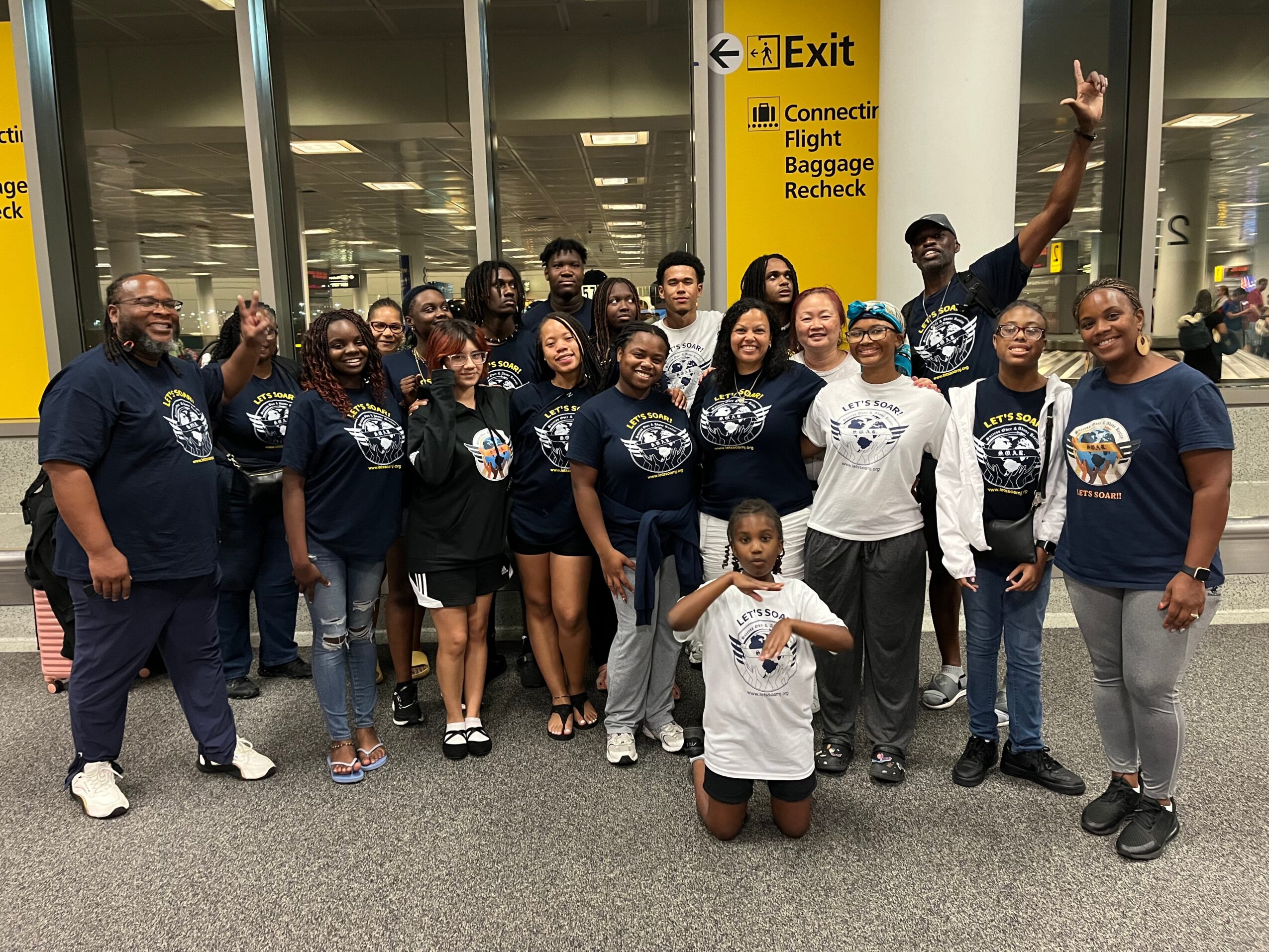 Group of people standing together at an airport terminal, some smiling and waving, with a child in front holding a paper.