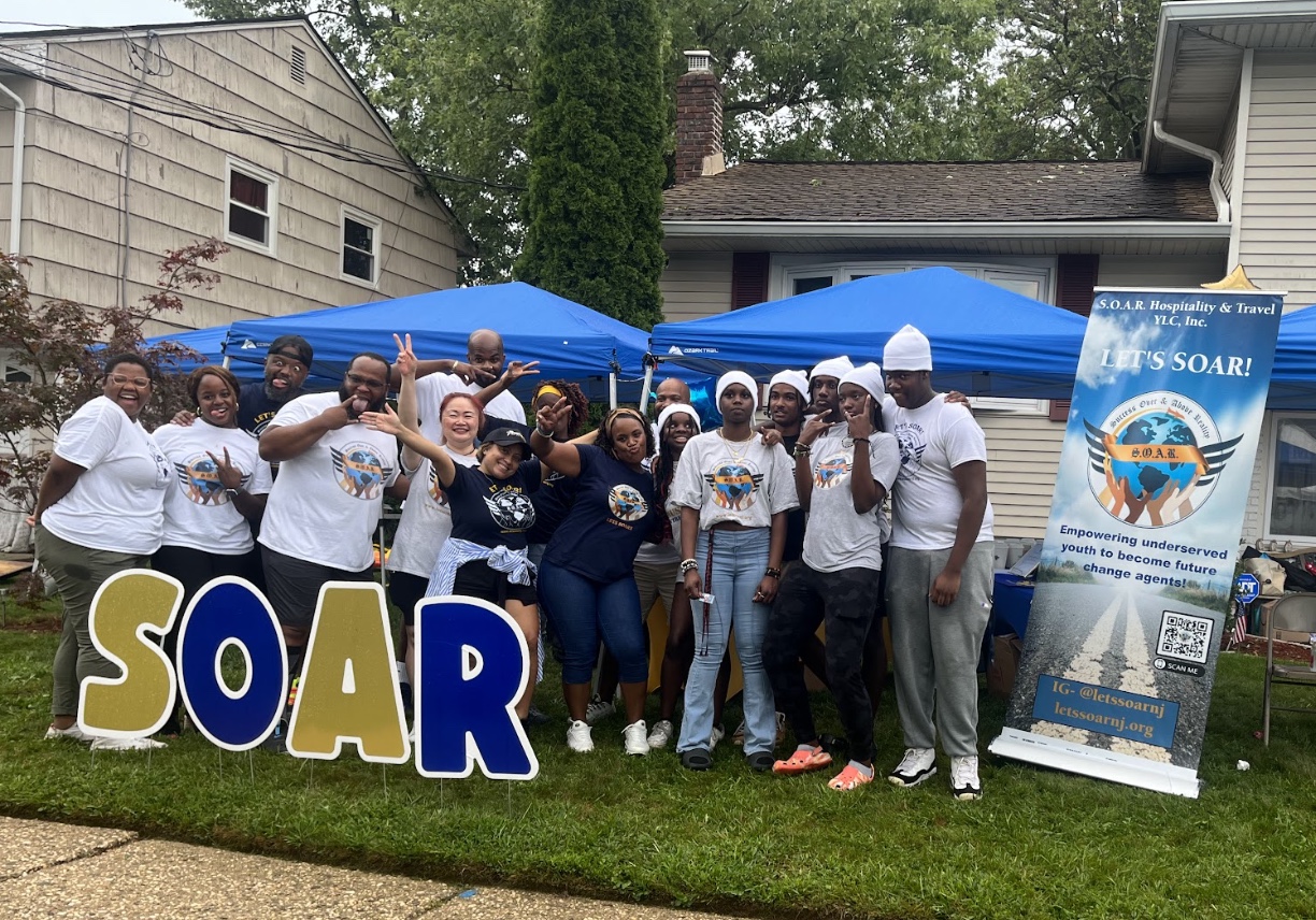 Group of people standing outdoors with blue tents, holding signs, and smiling, in front of a house and trees.