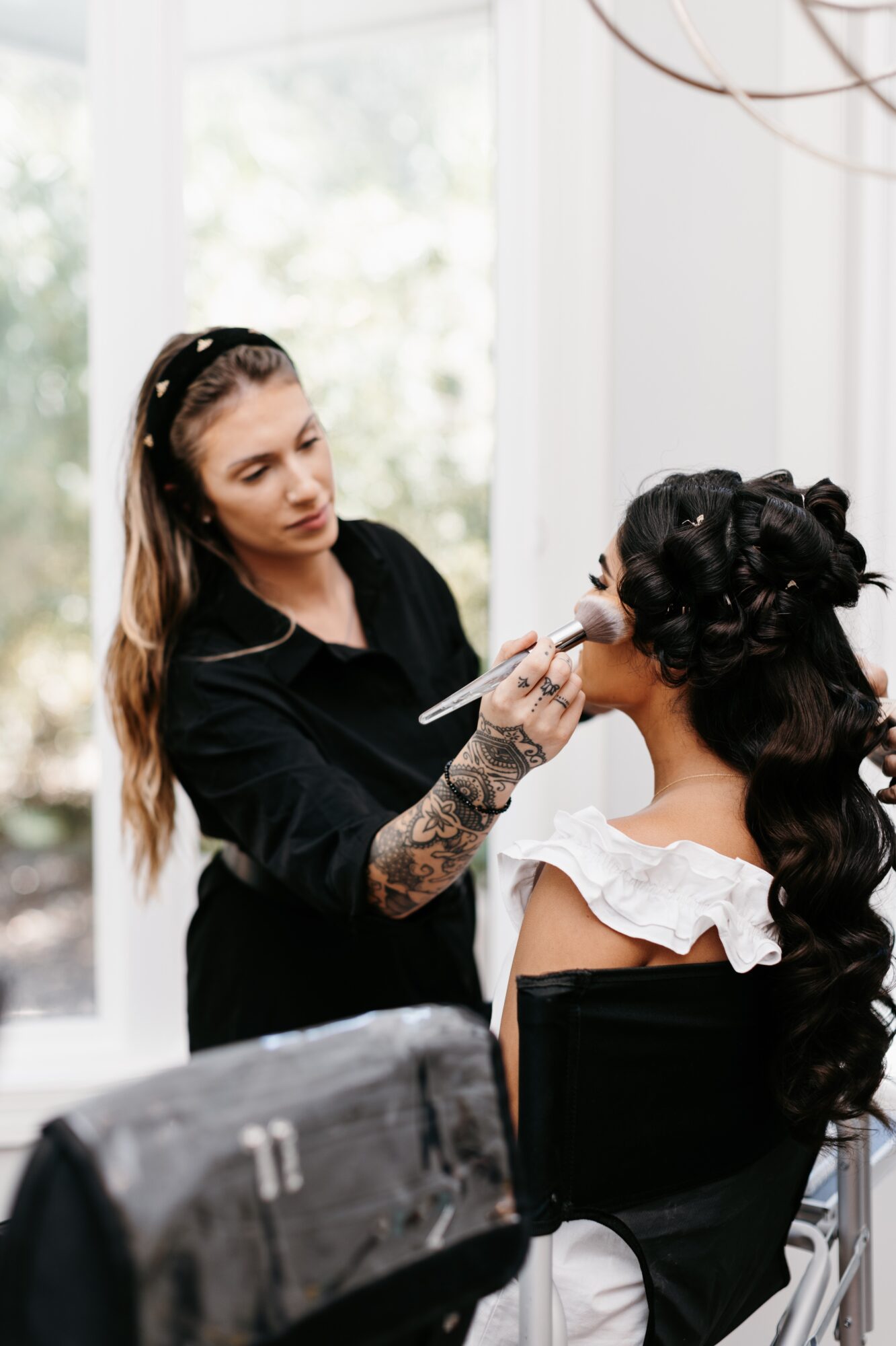 Makeup artist applying makeup to a woman with long, dark, curly hair in a bright room with large windows.