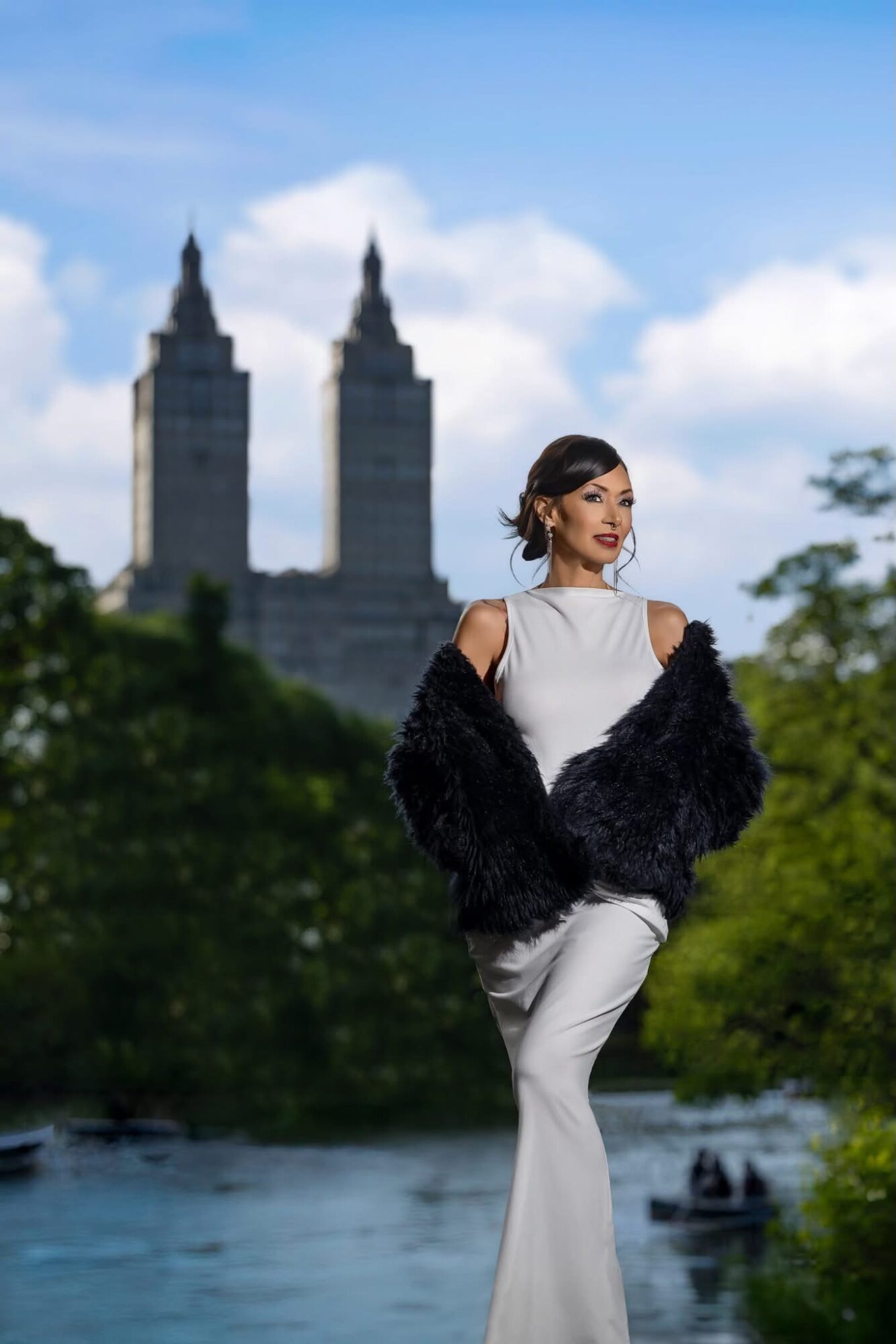 Woman in white dress and black fur coat standing outdoors with trees and a river, city skyline with tall buildings in background.