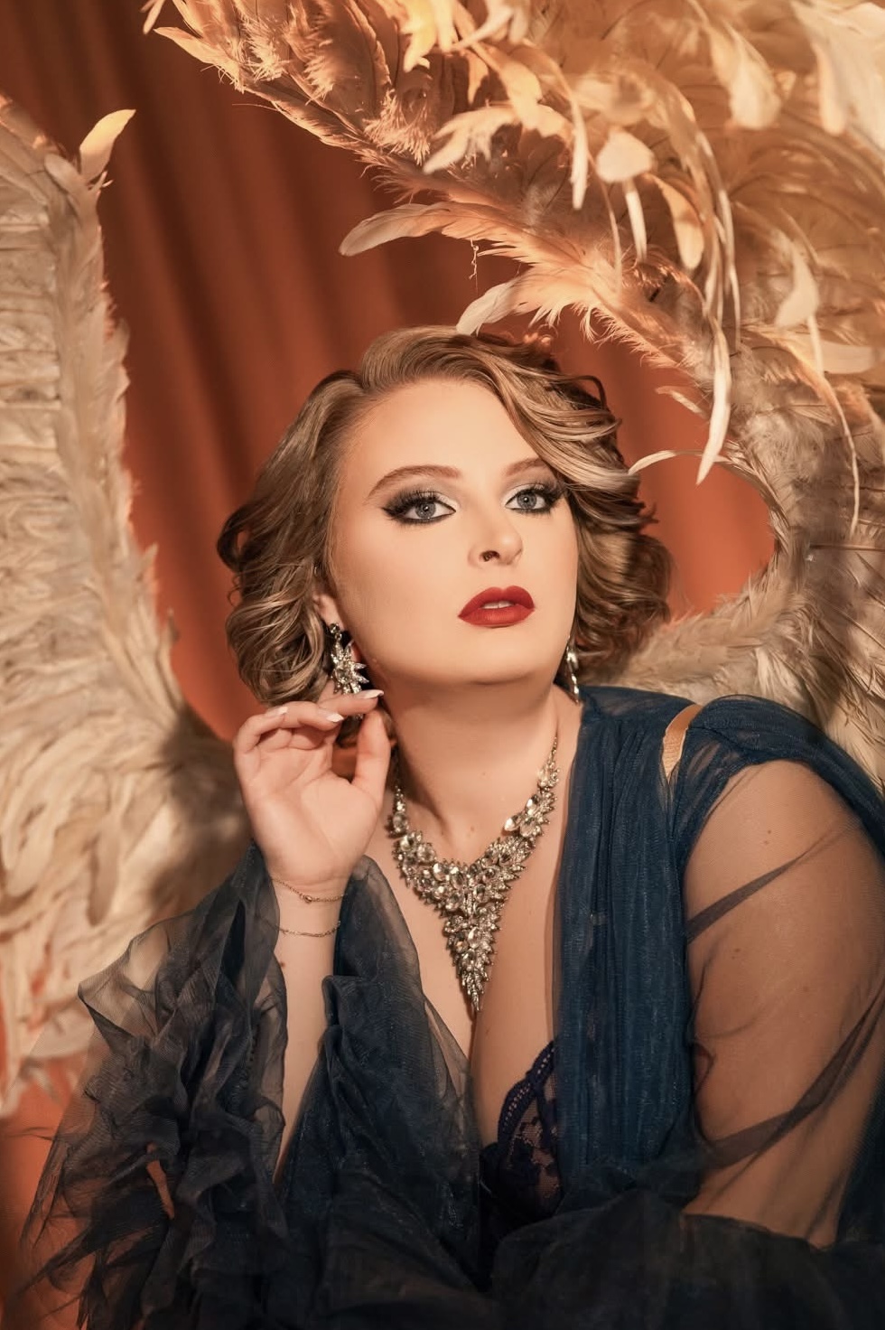 Woman with short wavy hair, red lipstick, wearing a dark blue dress and jewelry, posing with a feathered background.