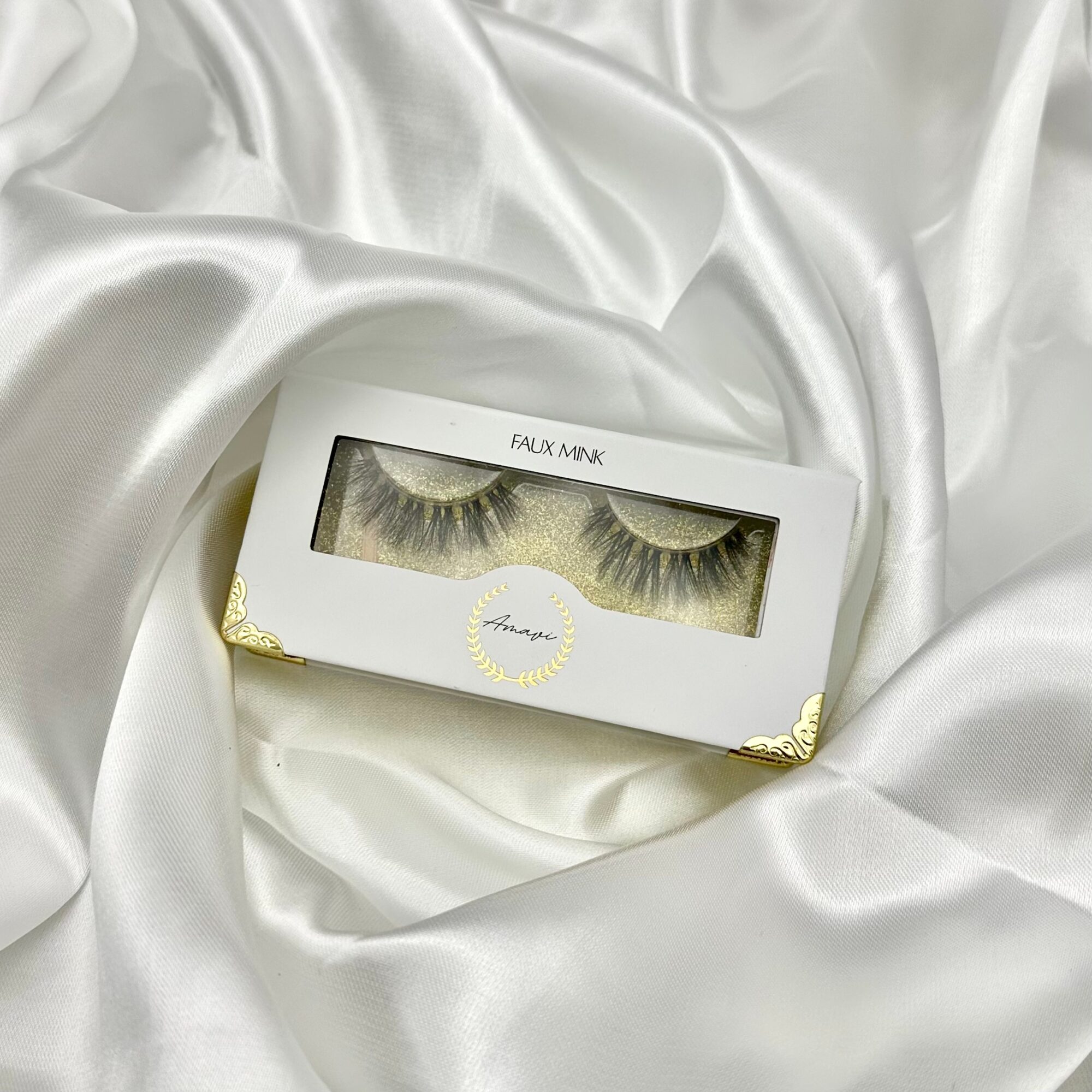 Box of false eyelashes on white satin fabric, gold accents on packaging, clear window showing eyelashes, elegant presentation.