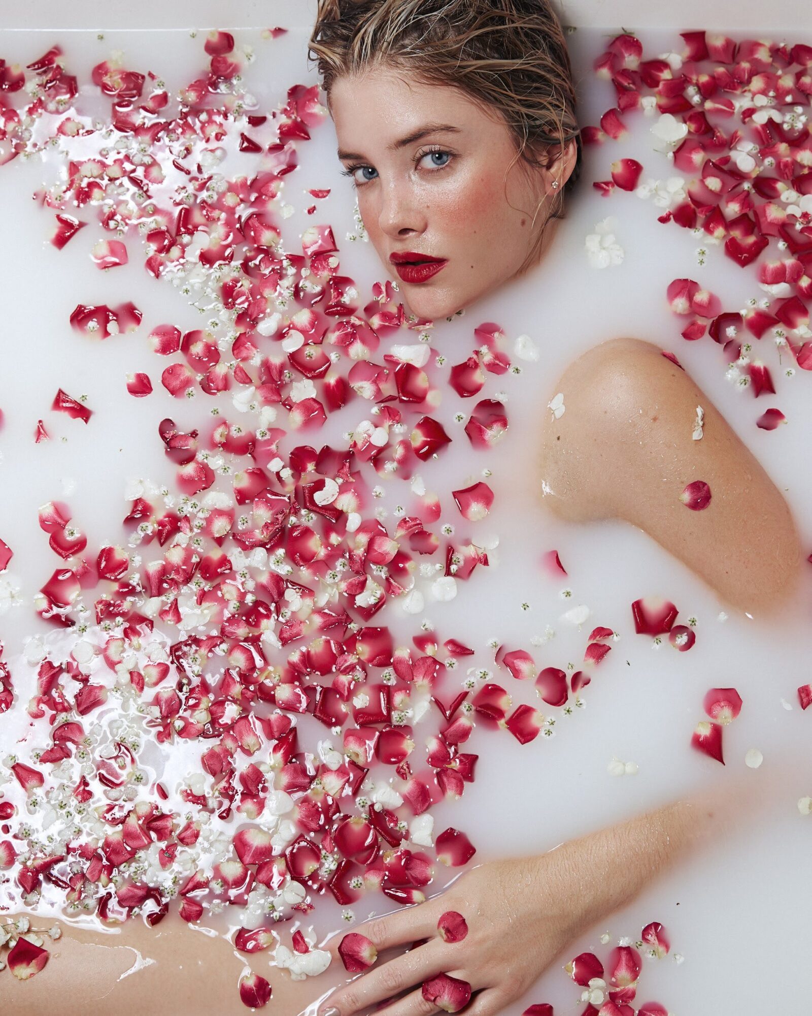 Young woman with wet hair surrounded by pink rose petals in a milky bath, looking at camera with neutral expression.