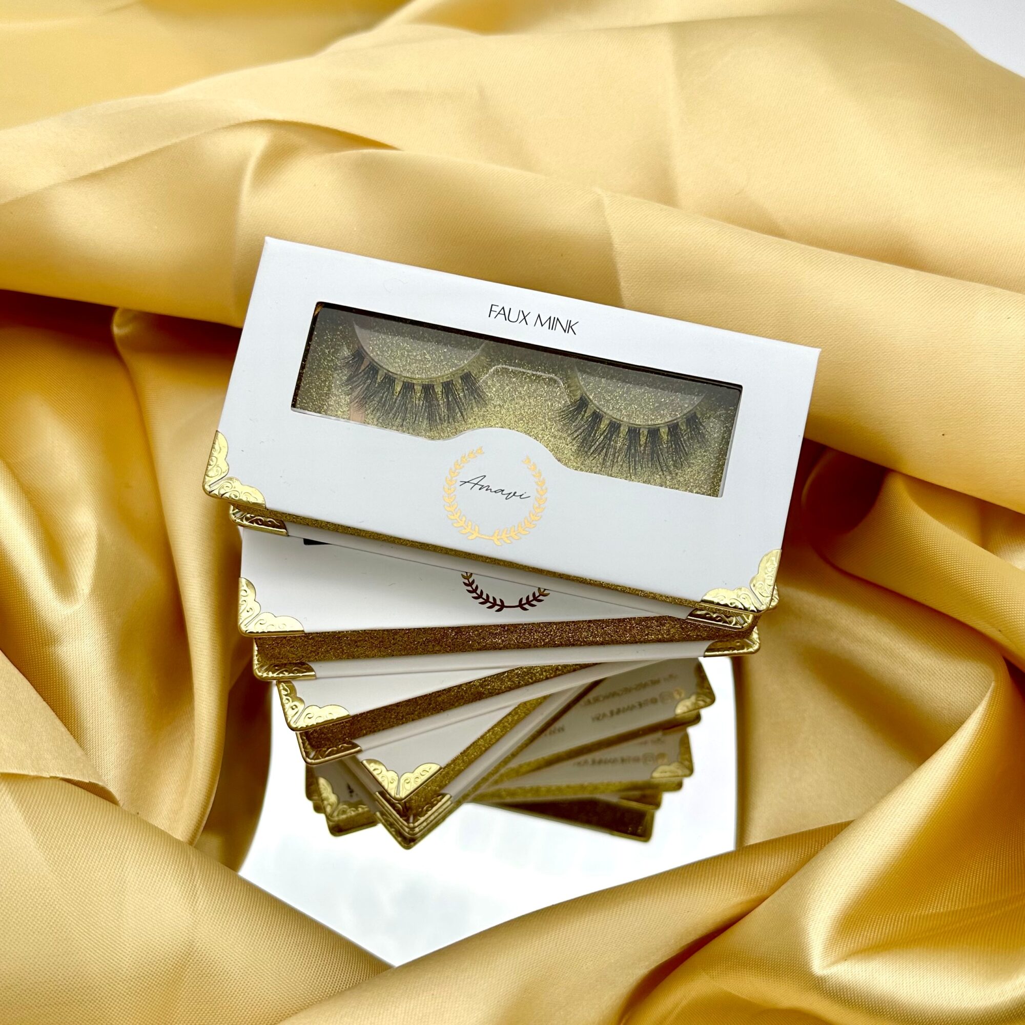 Box of false eyelashes on gold fabric with mirror reflection, gold accents, and a white label reading 'FAUX MINK'.