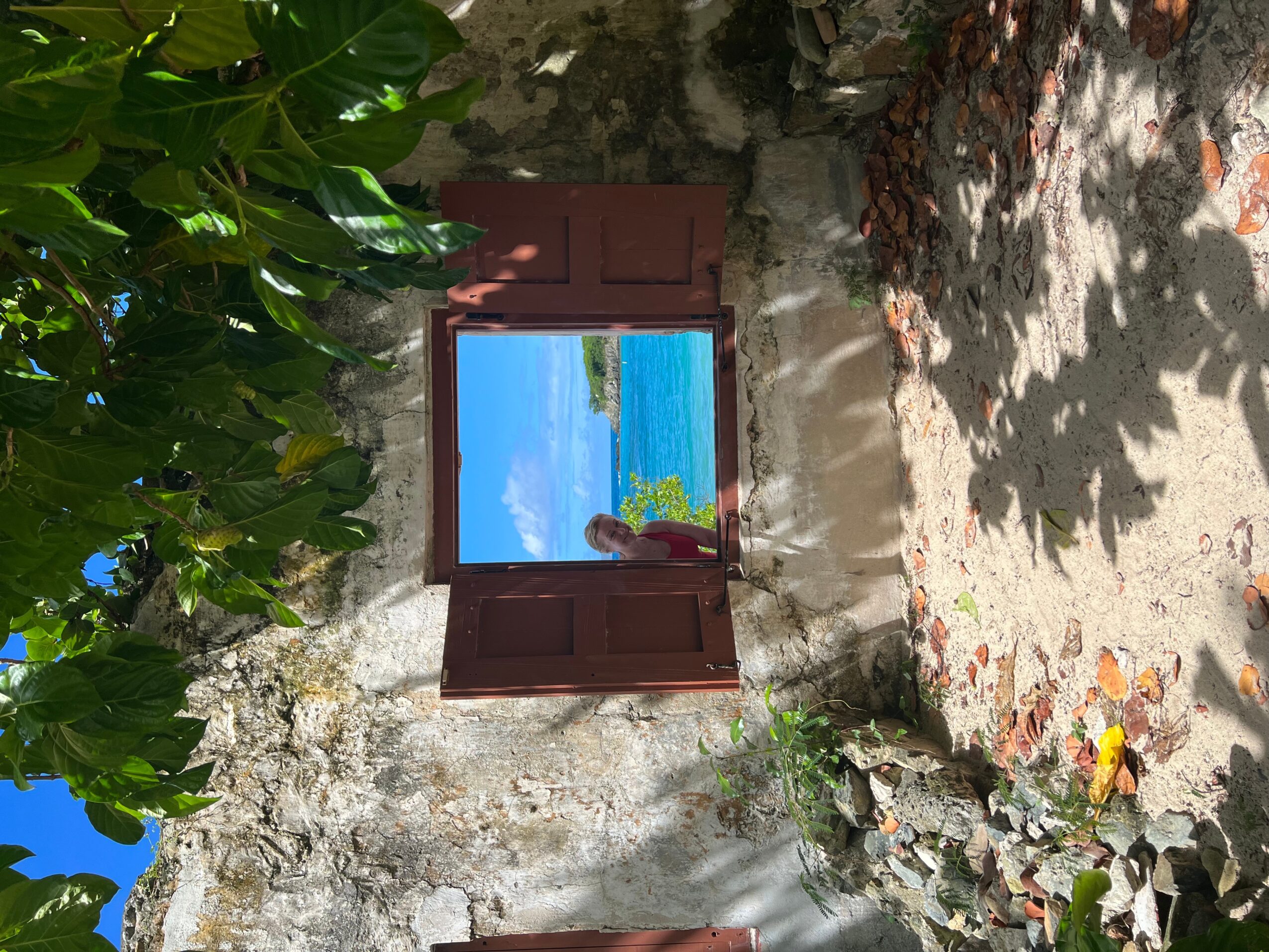 Open window with a view of the ocean and sky, surrounded by stone wall and greenery, sunlight casting shadows.