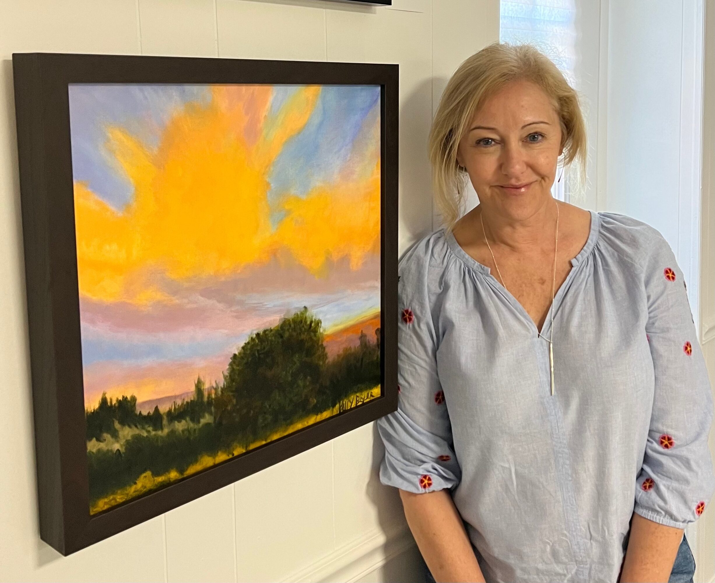 Woman with blonde hair standing next to a colorful landscape painting, smiling, in a bright room.