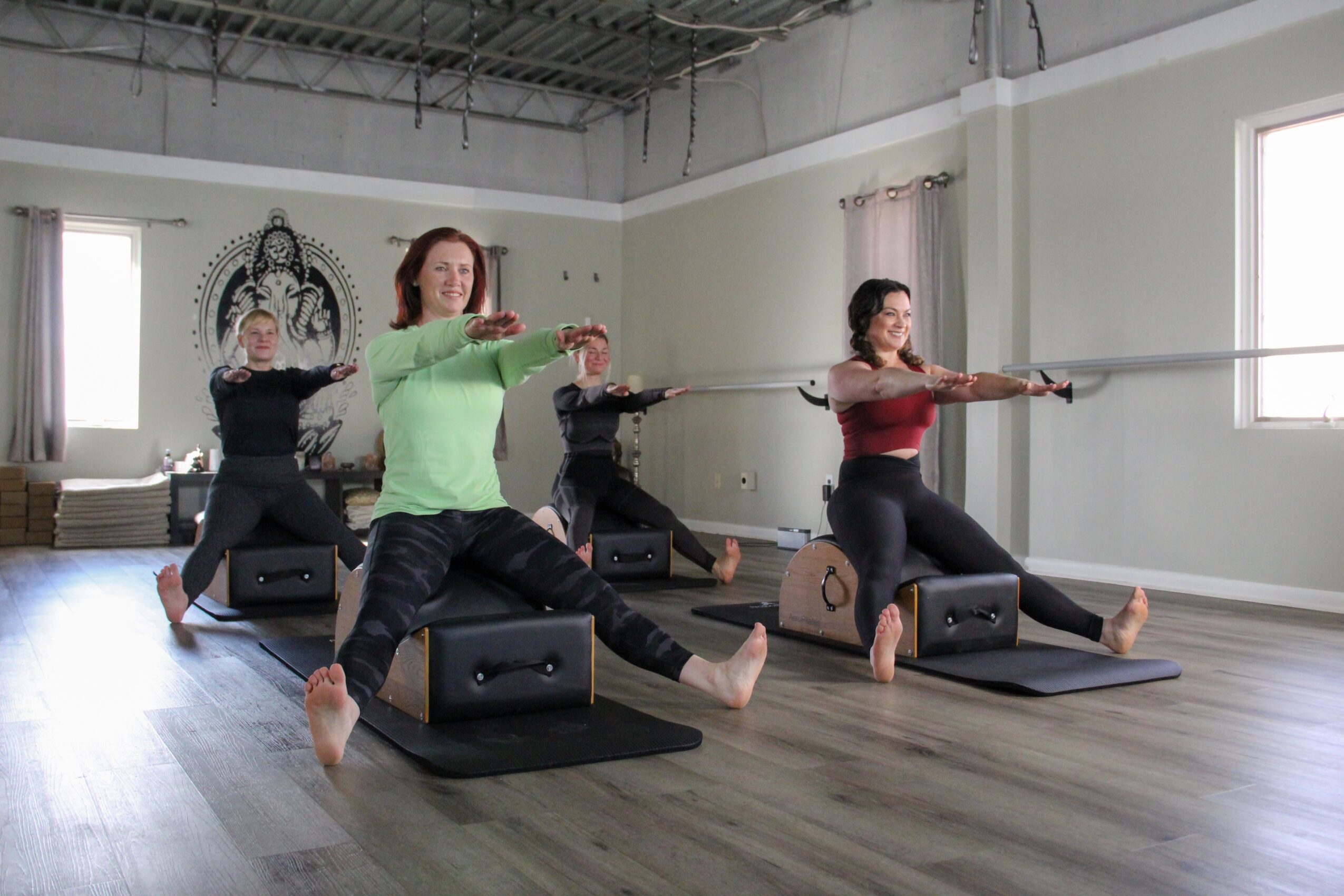 Four women participating in a fitness class using rowing machines in a bright studio with large windows.