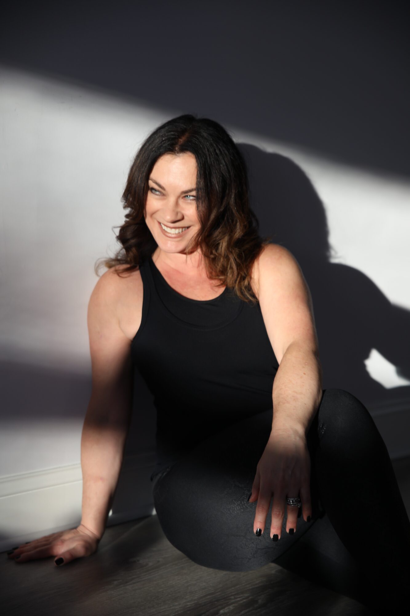 Woman with shoulder-length dark hair smiling, sitting on the floor in a black sleeveless top, with a shadow cast on the wall behind her.