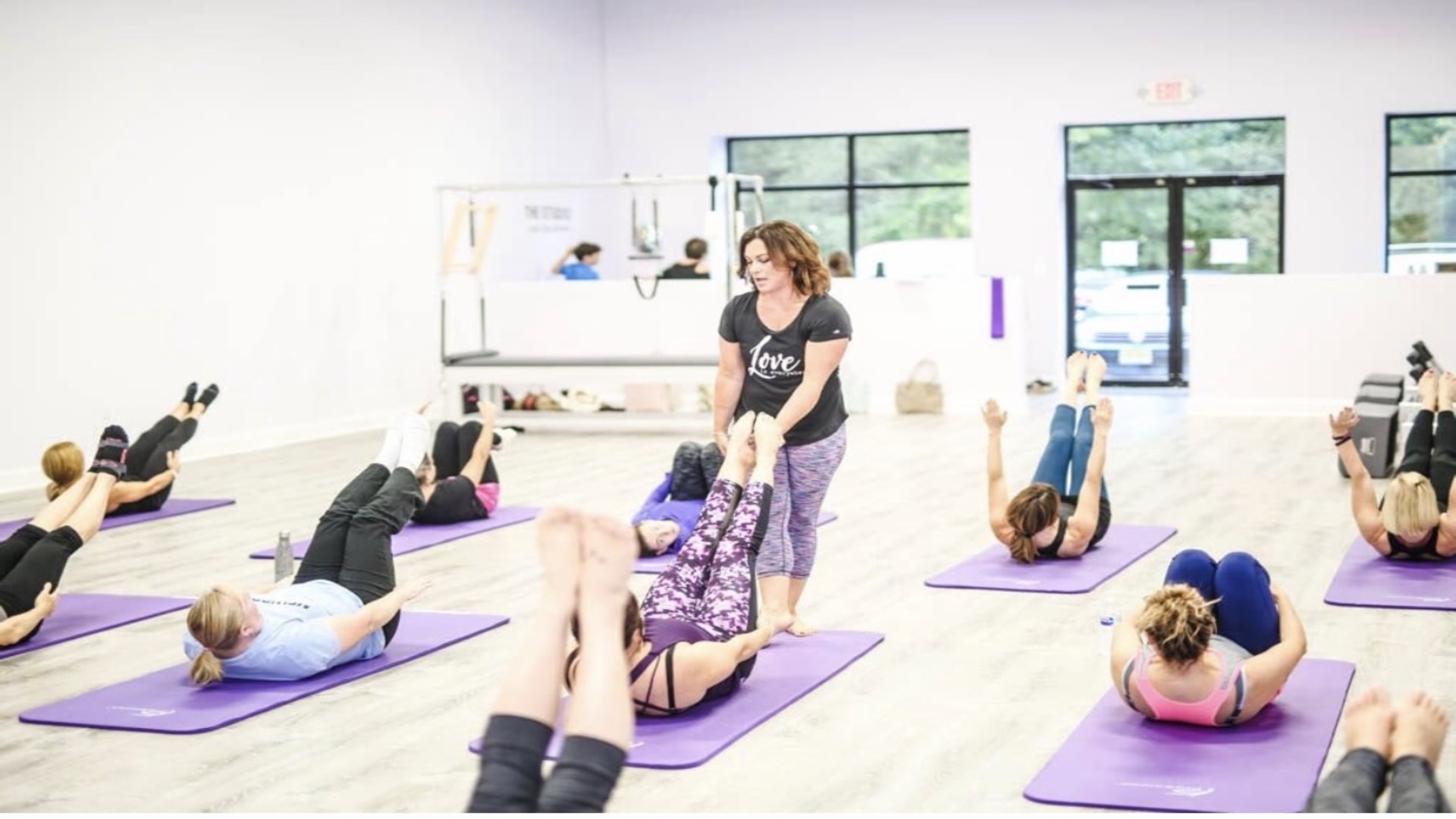 Group of people participating in a fitness class lying on purple mats, instructor standing in the center, bright room with large windows.