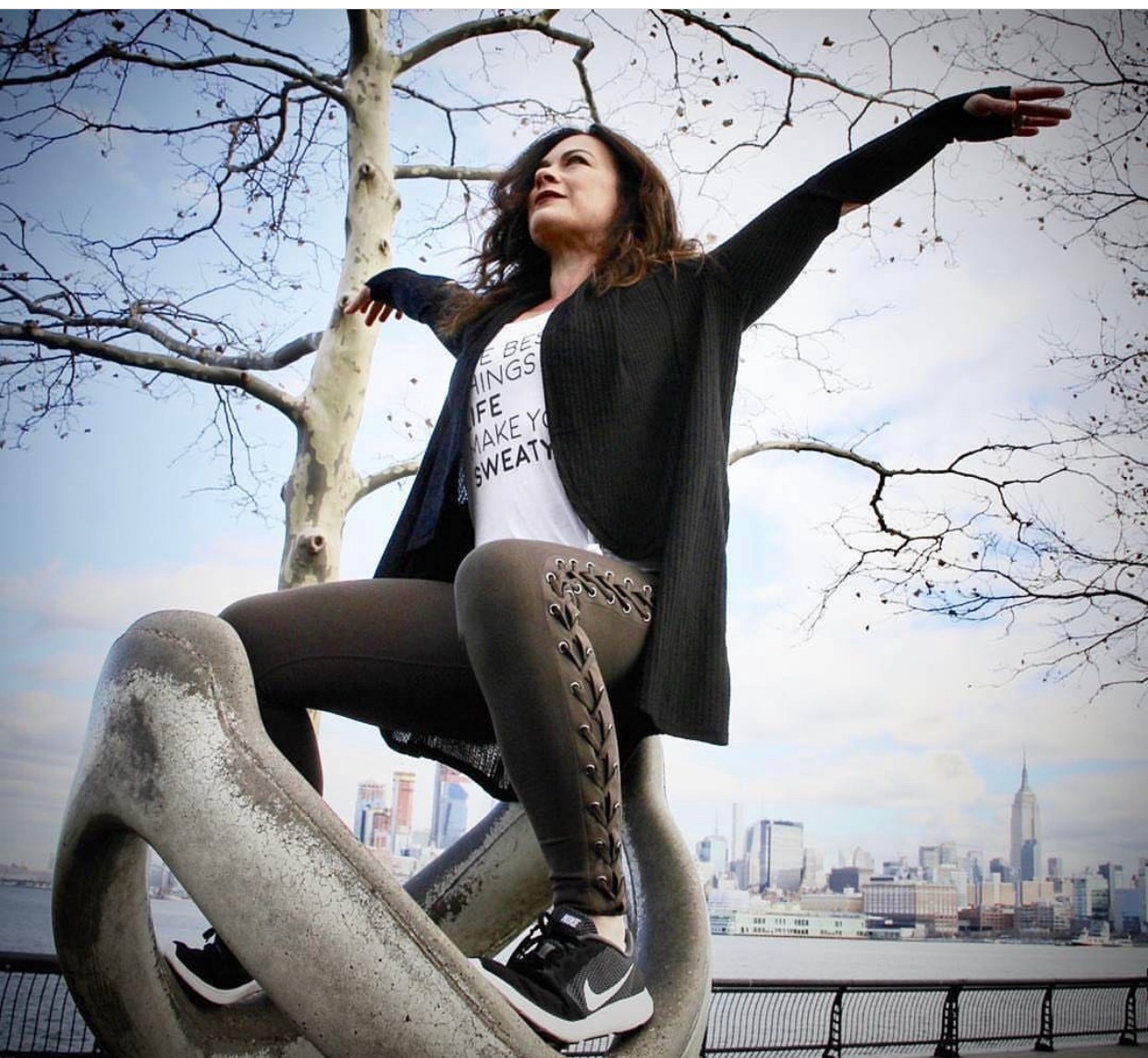 Woman standing on a sculpture with arms outstretched, city skyline in background, leafless tree nearby, cloudy sky.