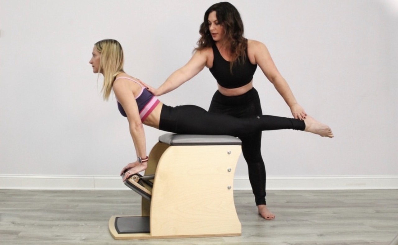 Woman assisting a girl with a stretching exercise on a padded bench, in a room with plain white walls.