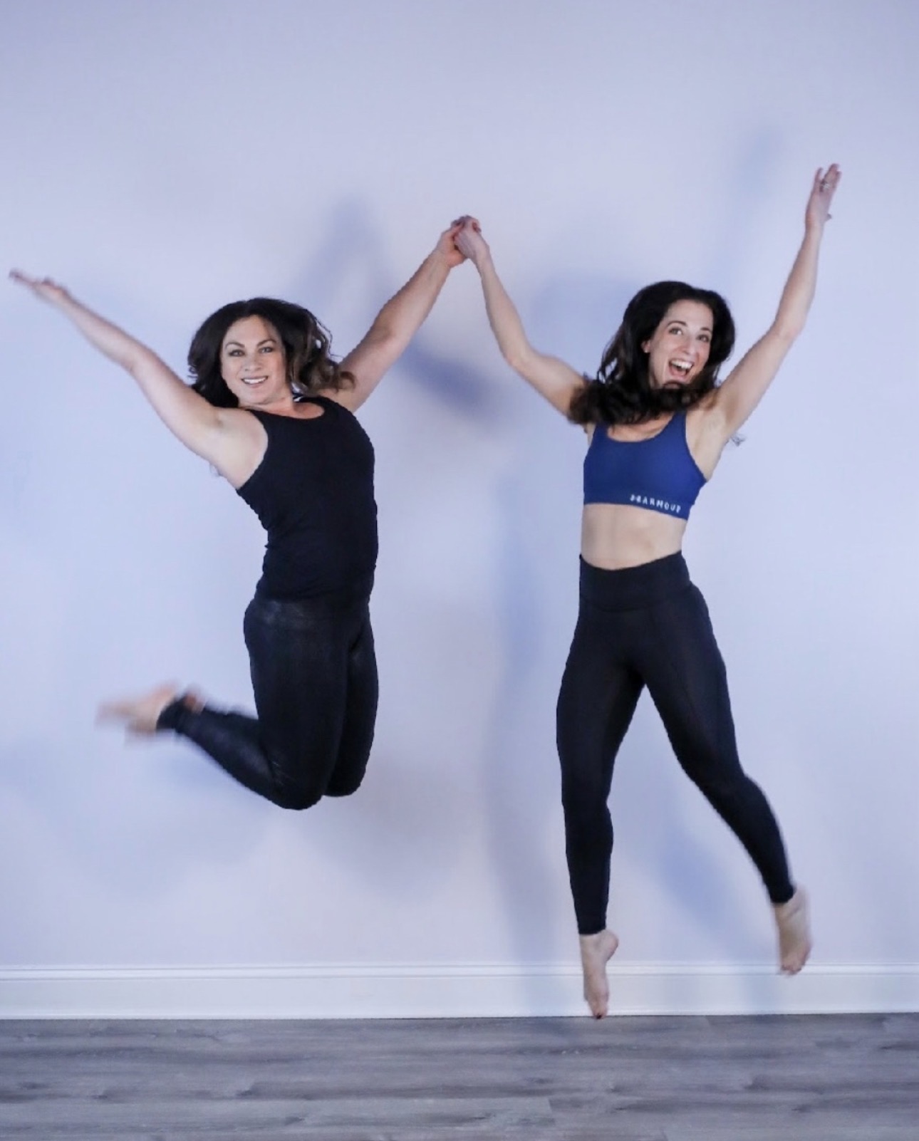 Two women jump in the air holding hands, smiling, against a plain light background.