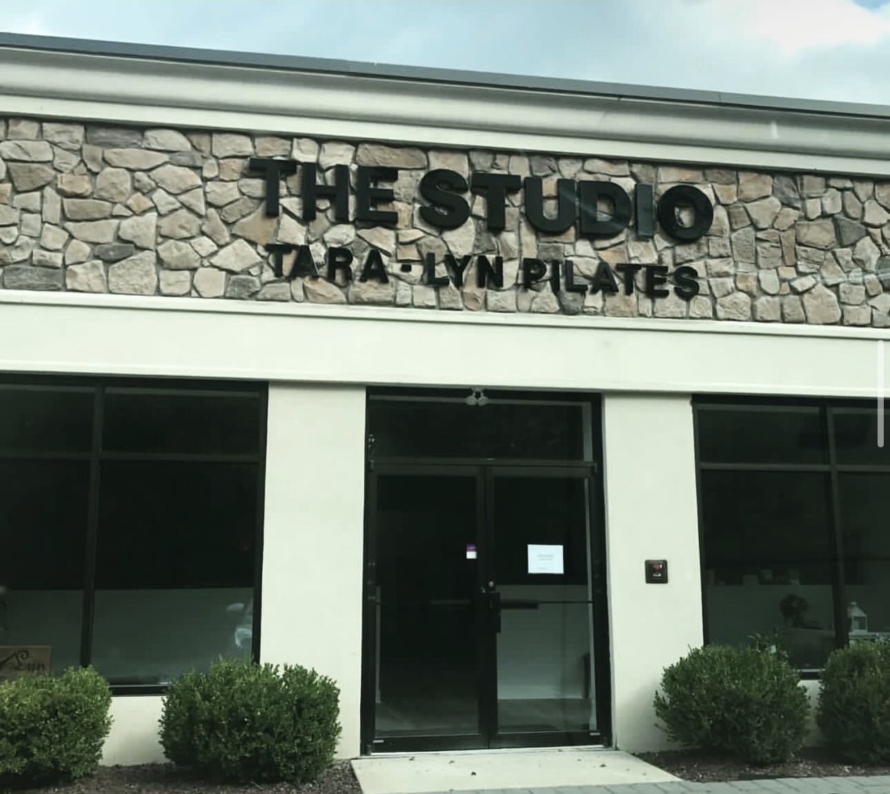 Storefront with stone facade and large windows, sign reads 'THE STUDIO TARA LYN PILATES', two bushes in front.