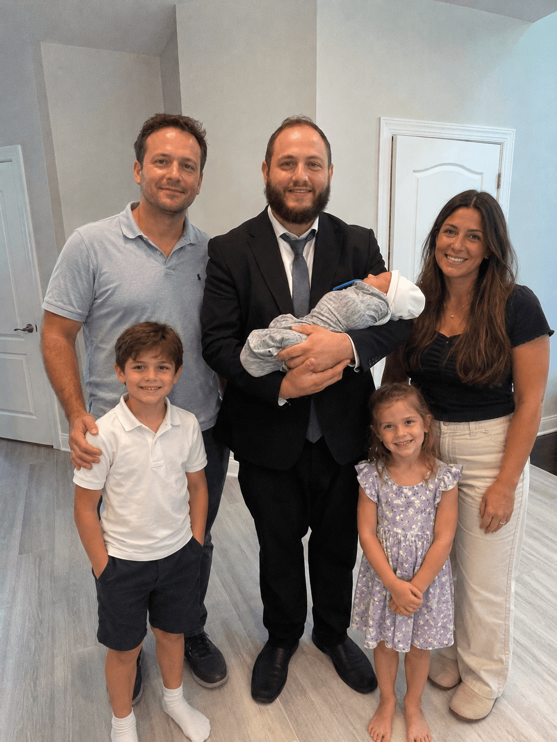 Group of six people, including a man in a suit holding a baby, two children, and three adults, standing indoors.