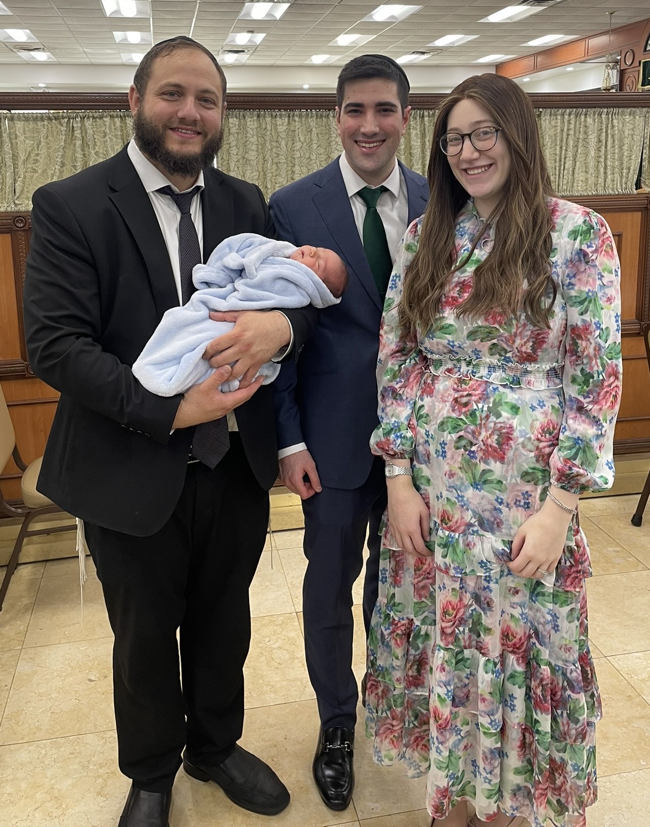Three people standing indoors, one holding a newborn wrapped in a blanket, all smiling, with curtains and chairs in background.