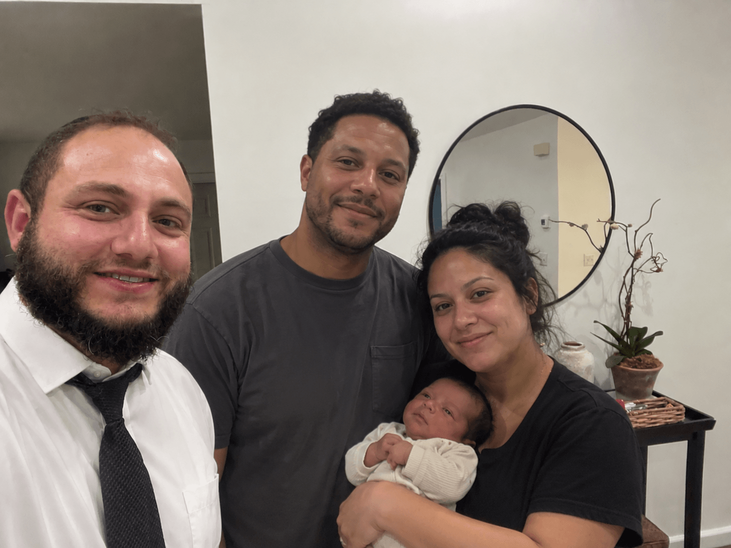 Group of four people, including a woman holding a baby, smiling indoors with a mirror and plant in the background.