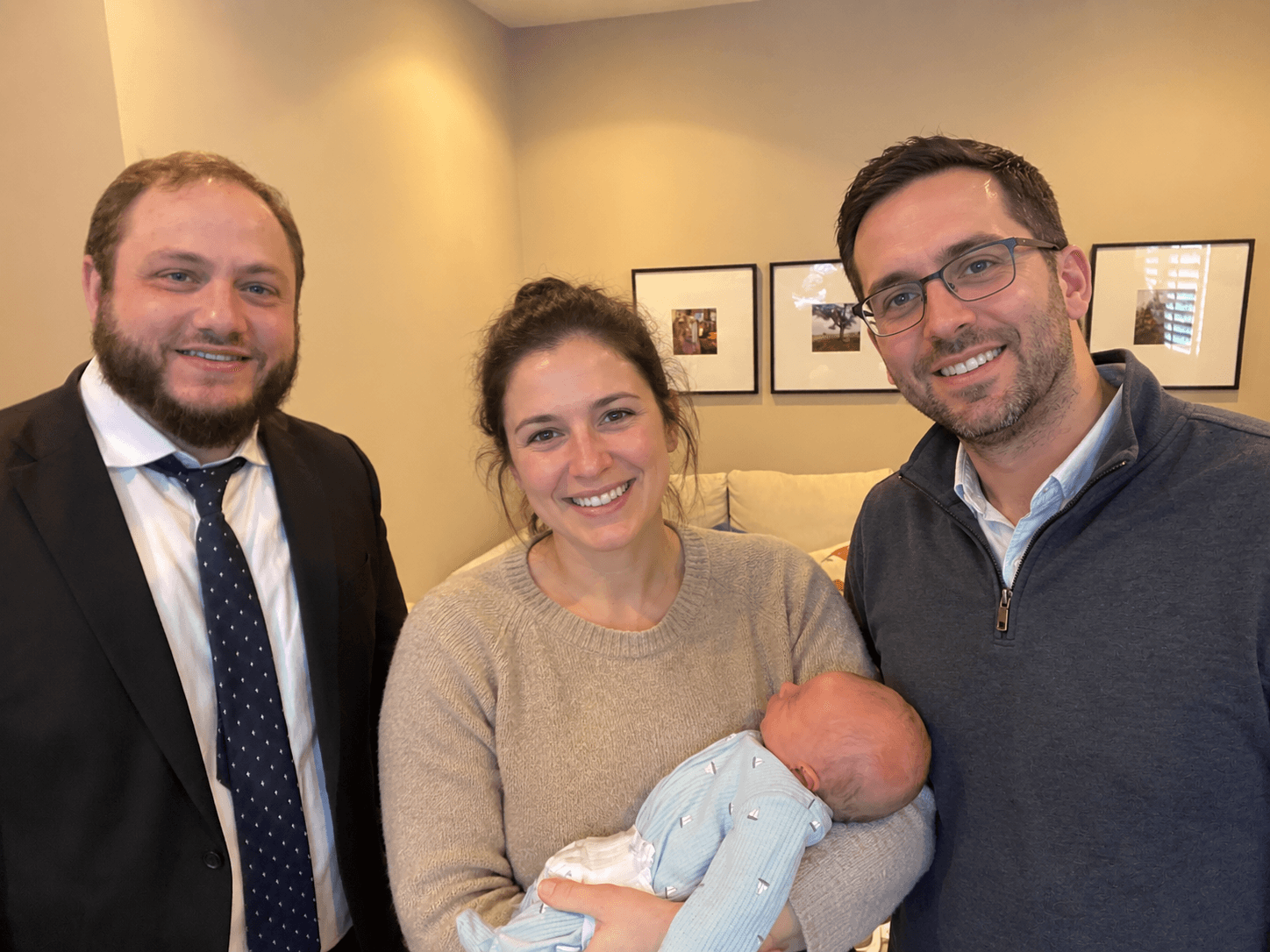 Three people, two men and a woman, holding a newborn baby indoors, smiling at the camera.