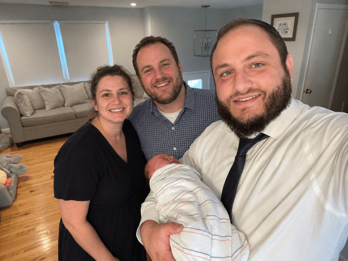 Three adults and a newborn baby in a living room, smiling for a selfie, with a sofa and framed pictures in the background.