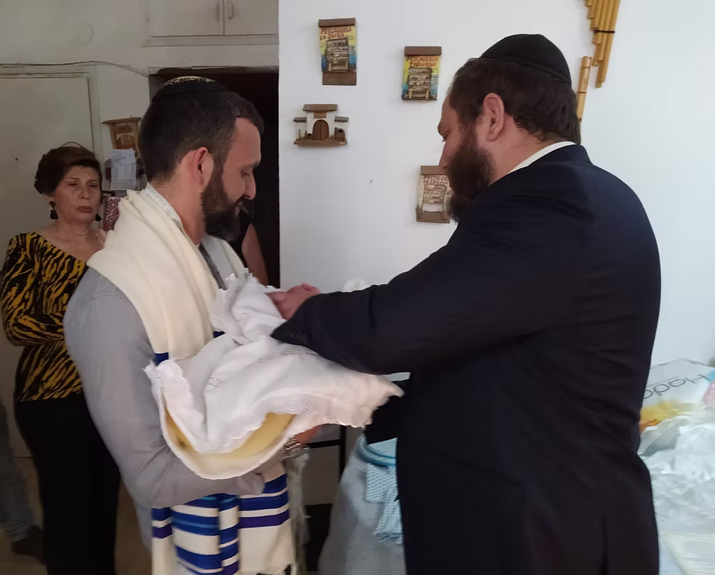 Two men holding a baby, one in a suit and the other in a tallit, with a woman observing in the background.
