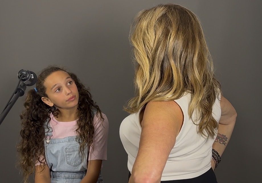 Two females facing each other, one with long curly hair and the other with shoulder-length wavy hair, in front of a gray background.