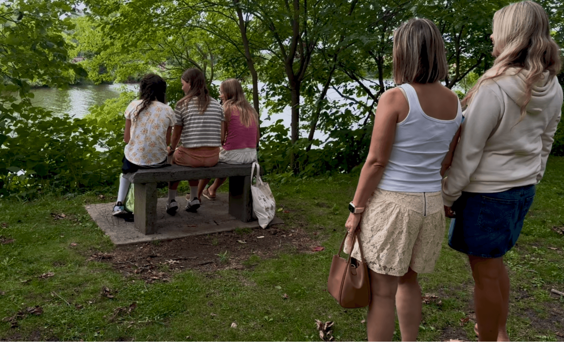 Four women by a lake, three sitting on a bench and two standing nearby, surrounded by trees and greenery.