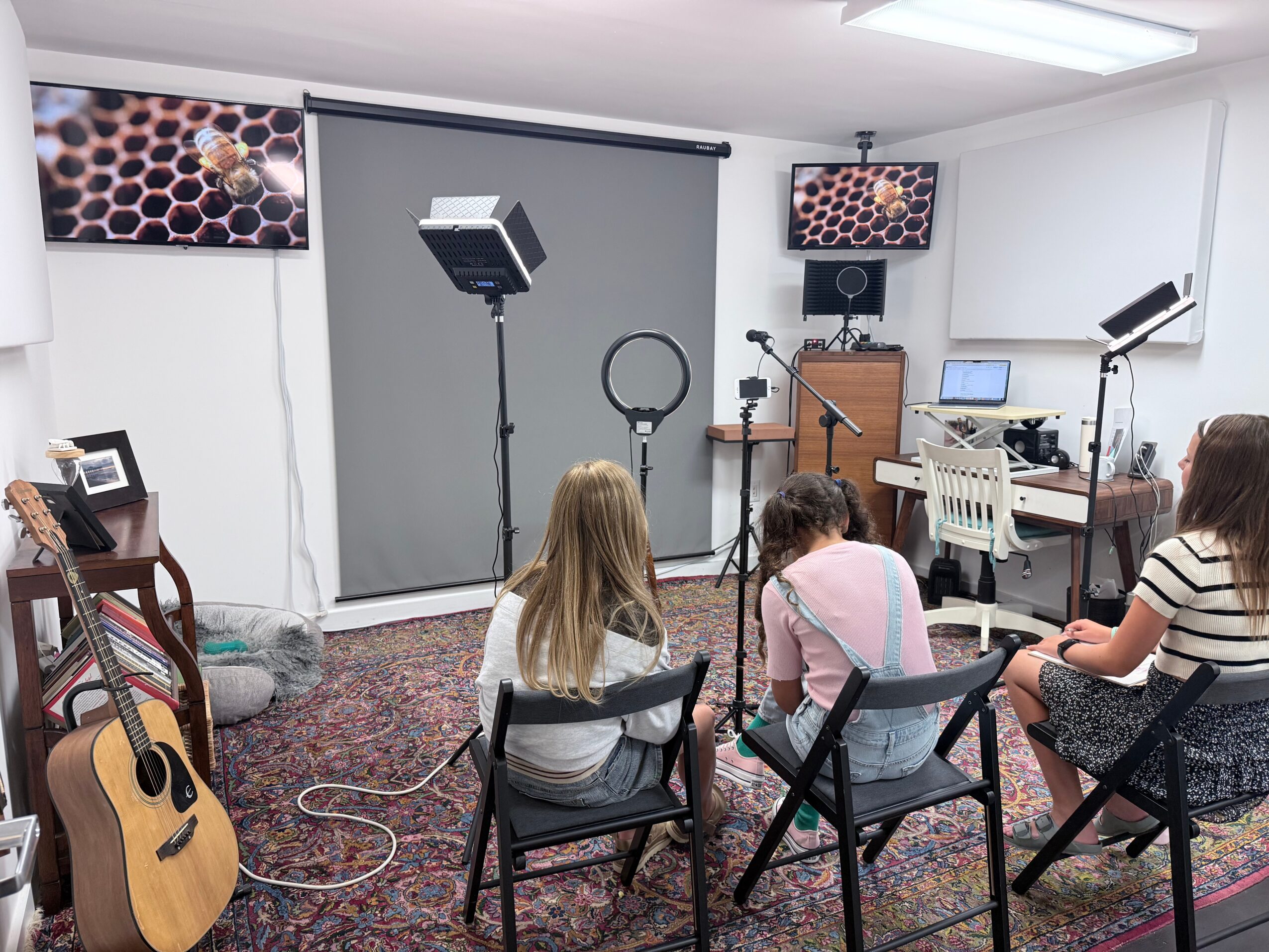 Three children sit on chairs in a room with recording equipment, a guitar, and a projector screen, facing a wall with screens.