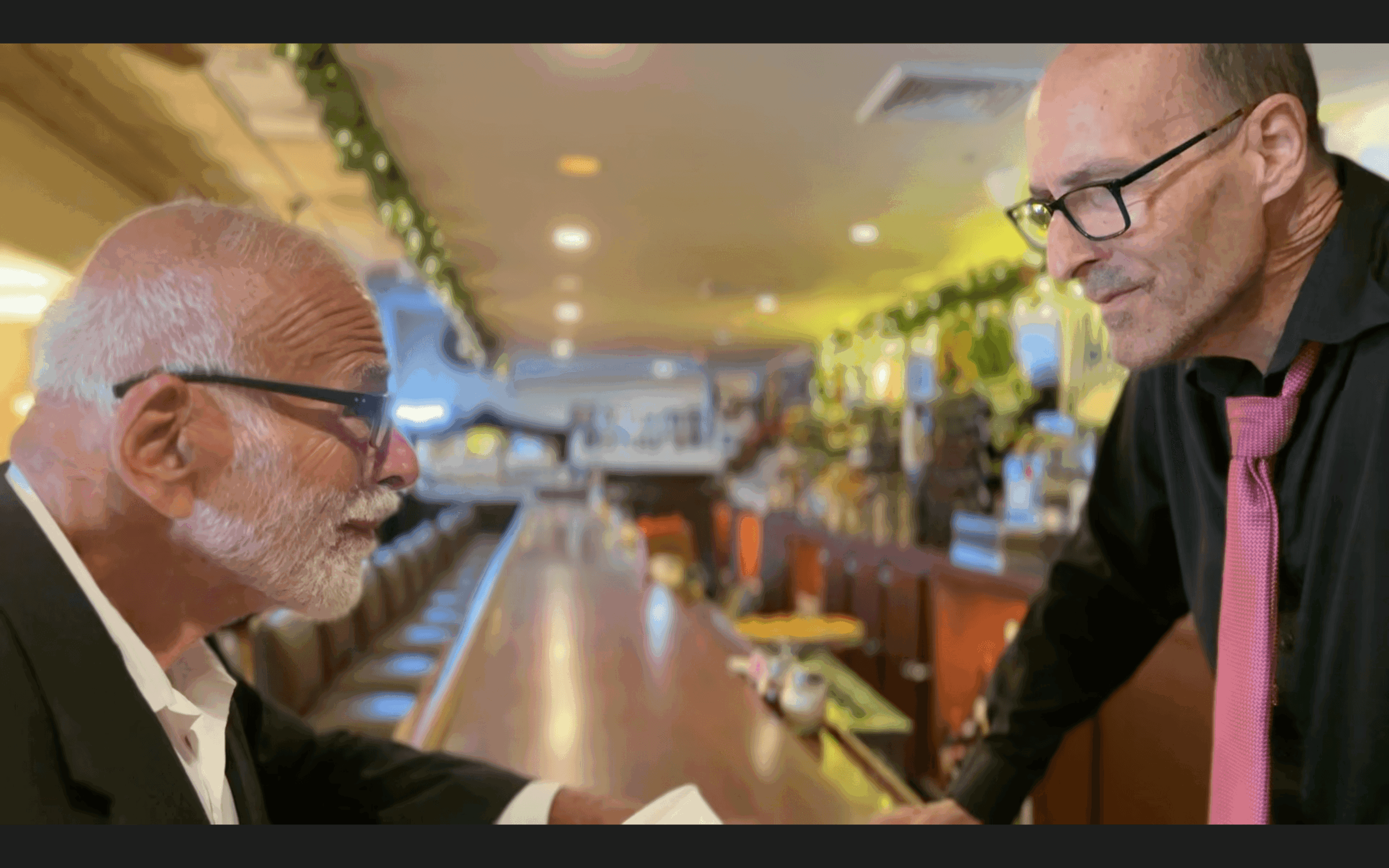 Two men converse at a bar counter, one seated and one standing, in a well-lit establishment with decorations.