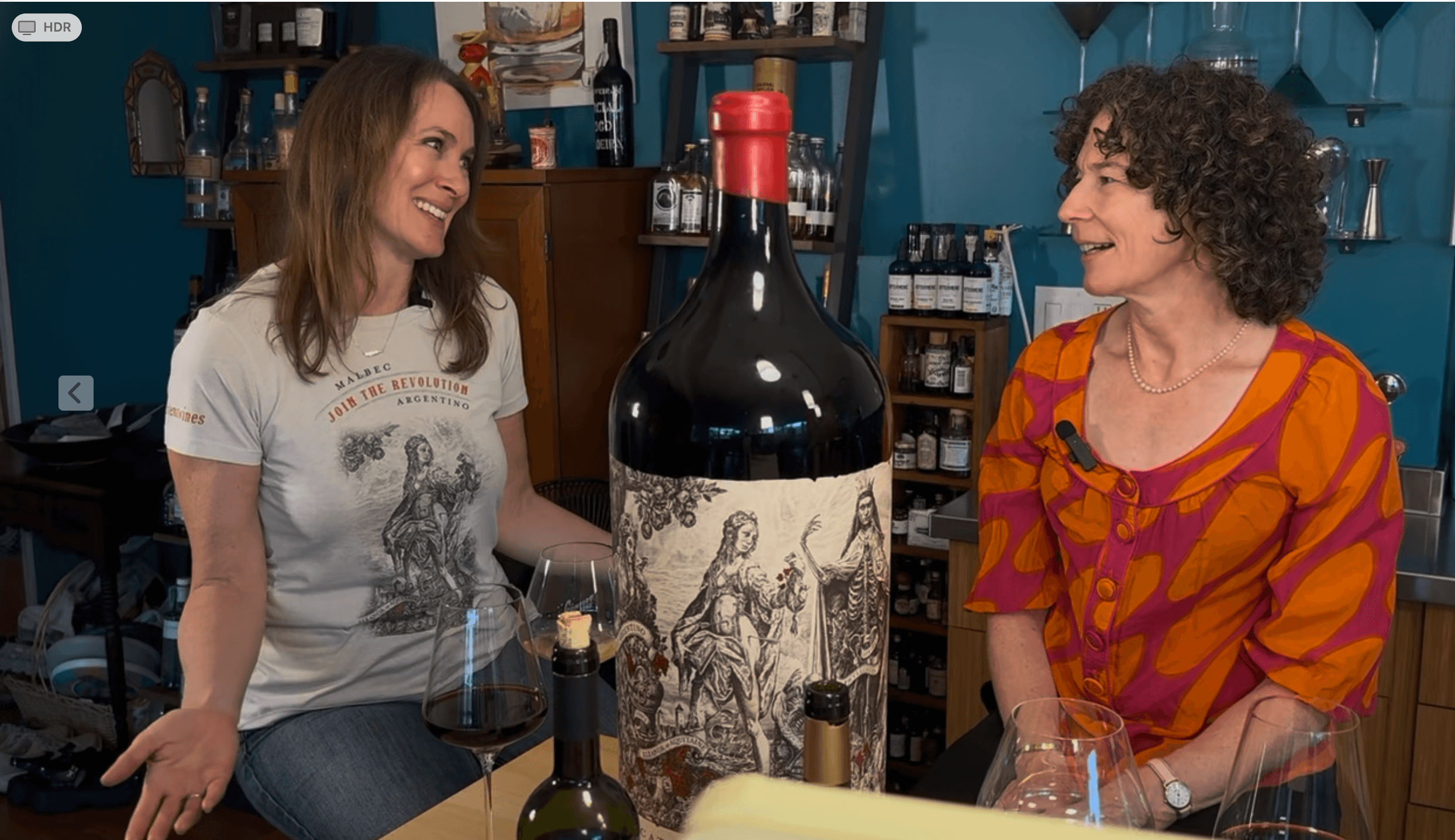 Two women smiling and talking in a wine shop with bottles and a large wine bottle illustration behind them.