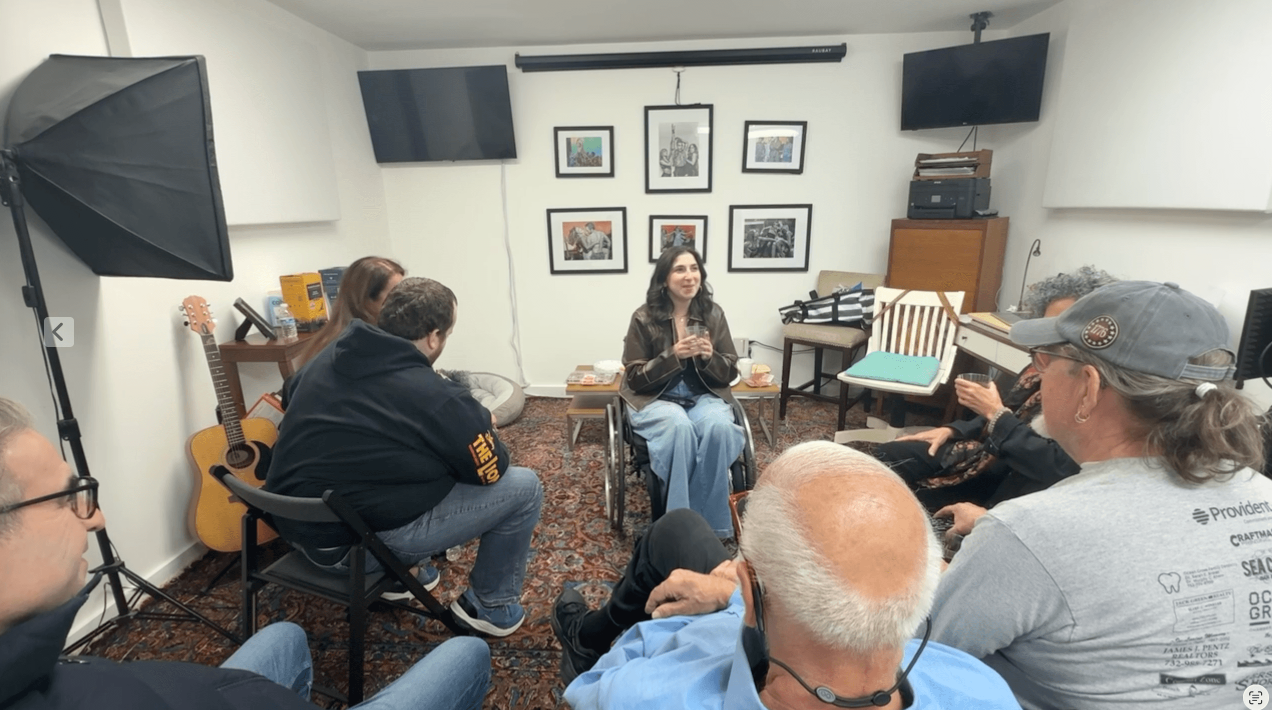 Group of people sitting in a room, listening to a woman speaking, with framed pictures on the wall and two TVs.