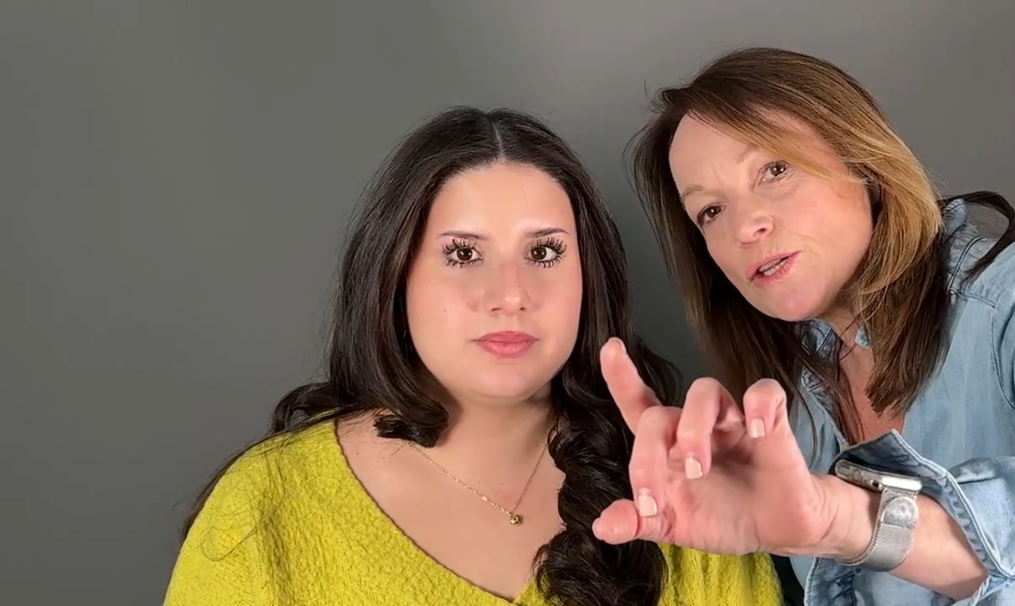 Two women with dark and light brown hair, one in a yellow top, pose against a gray background.