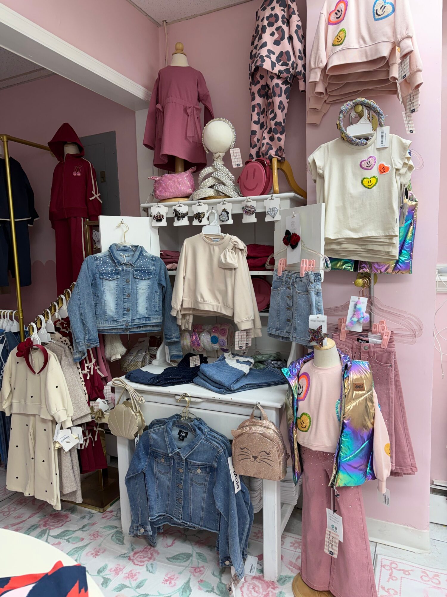 Display of children's clothing and accessories on pink shelves and a white rack, including dresses, shirts, and bags.