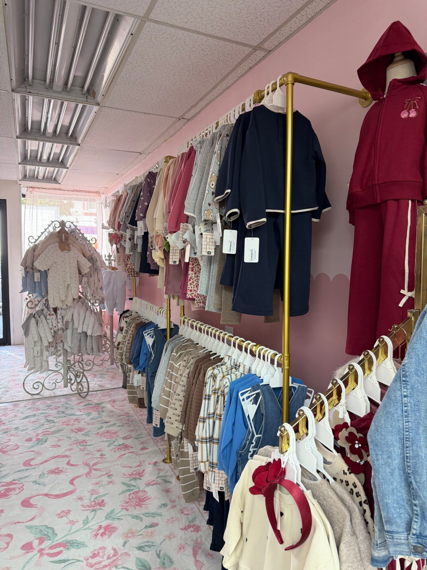 Clothing racks with jackets, shirts, and dresses in a store, pink wall, and floral carpet floor.