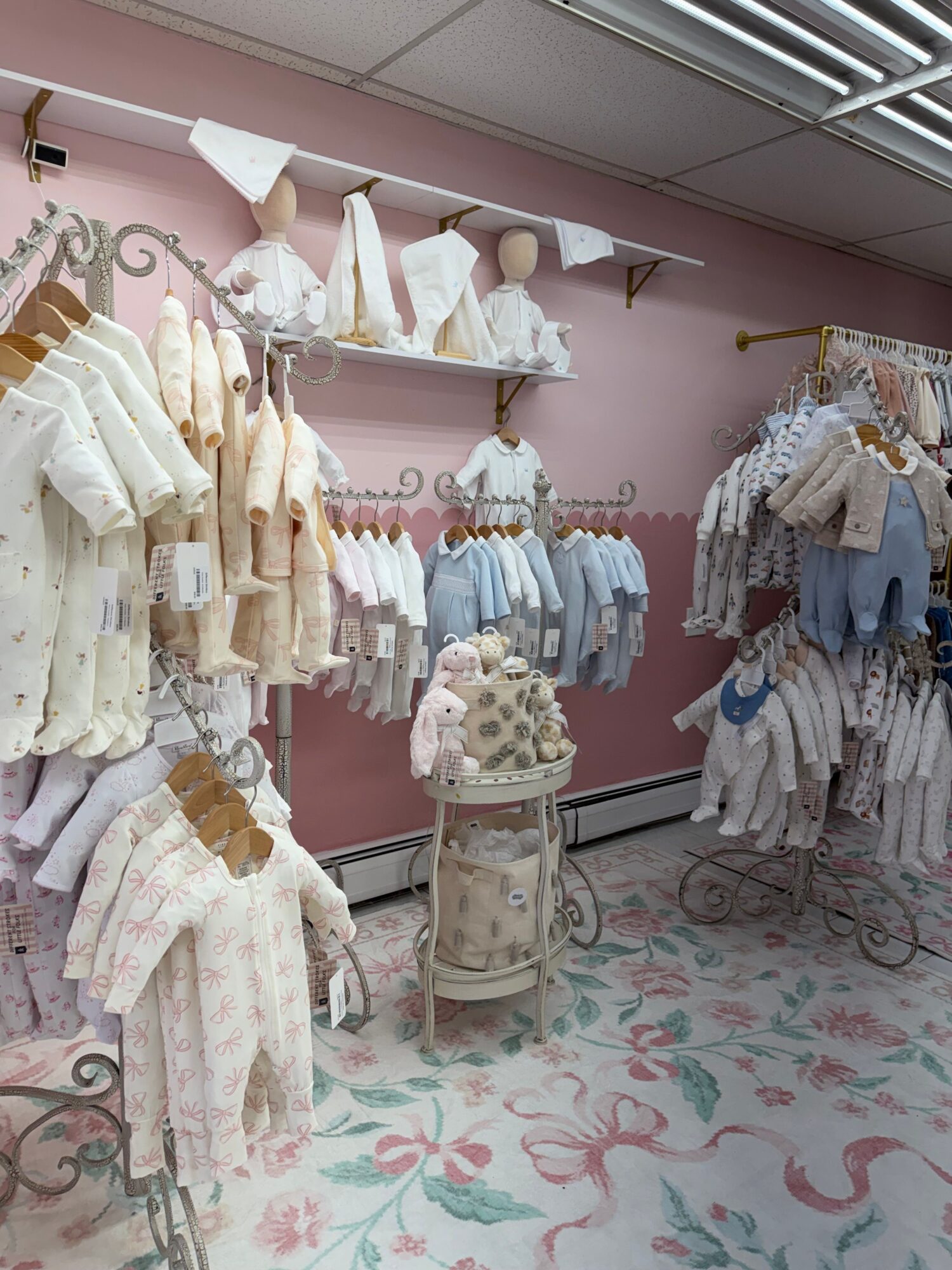 Display of baby clothes and accessories on tables and shelves in a pink room with floral carpet.