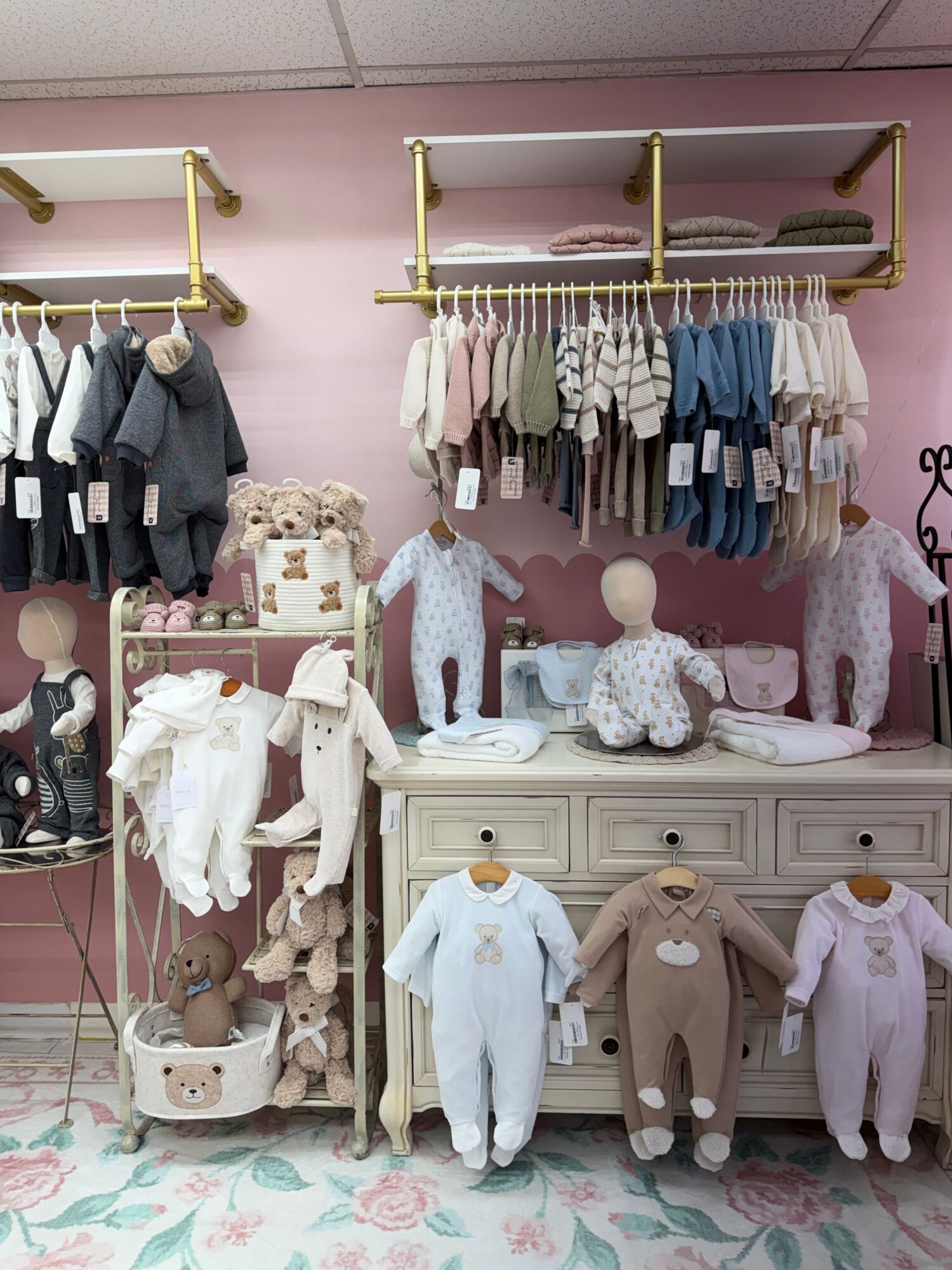 Display of clothing and accessories in a boutique with pink walls and floral carpet, including shirts, dresses, and mannequins.