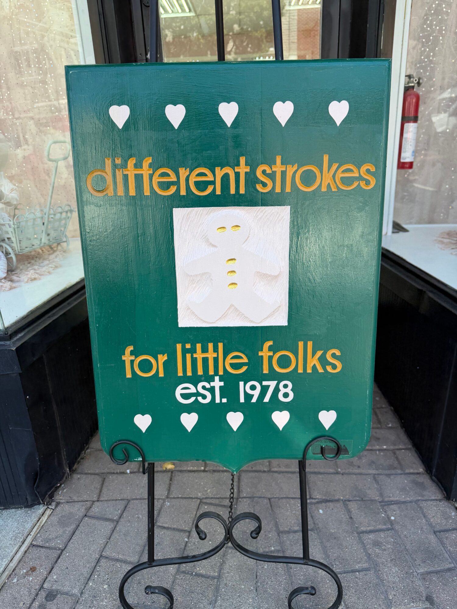 Green signboard with white hearts and text, standing on a sidewalk outside a building.