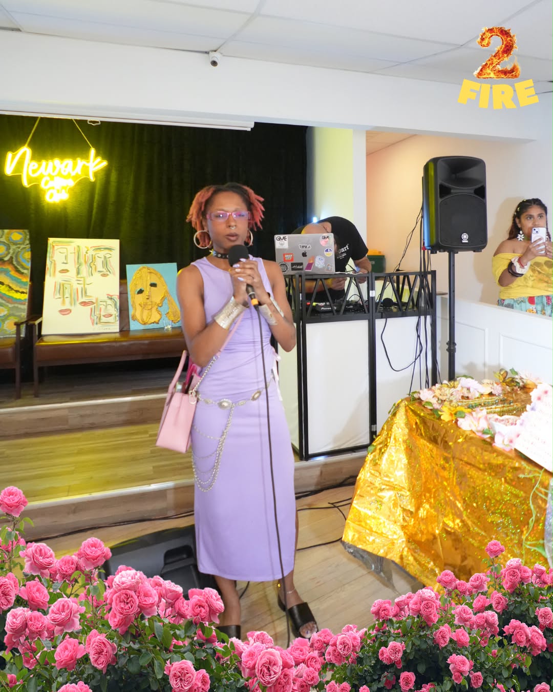 Woman in lavender dress holding microphone, standing near decorated table with pink flowers, in indoor event space.