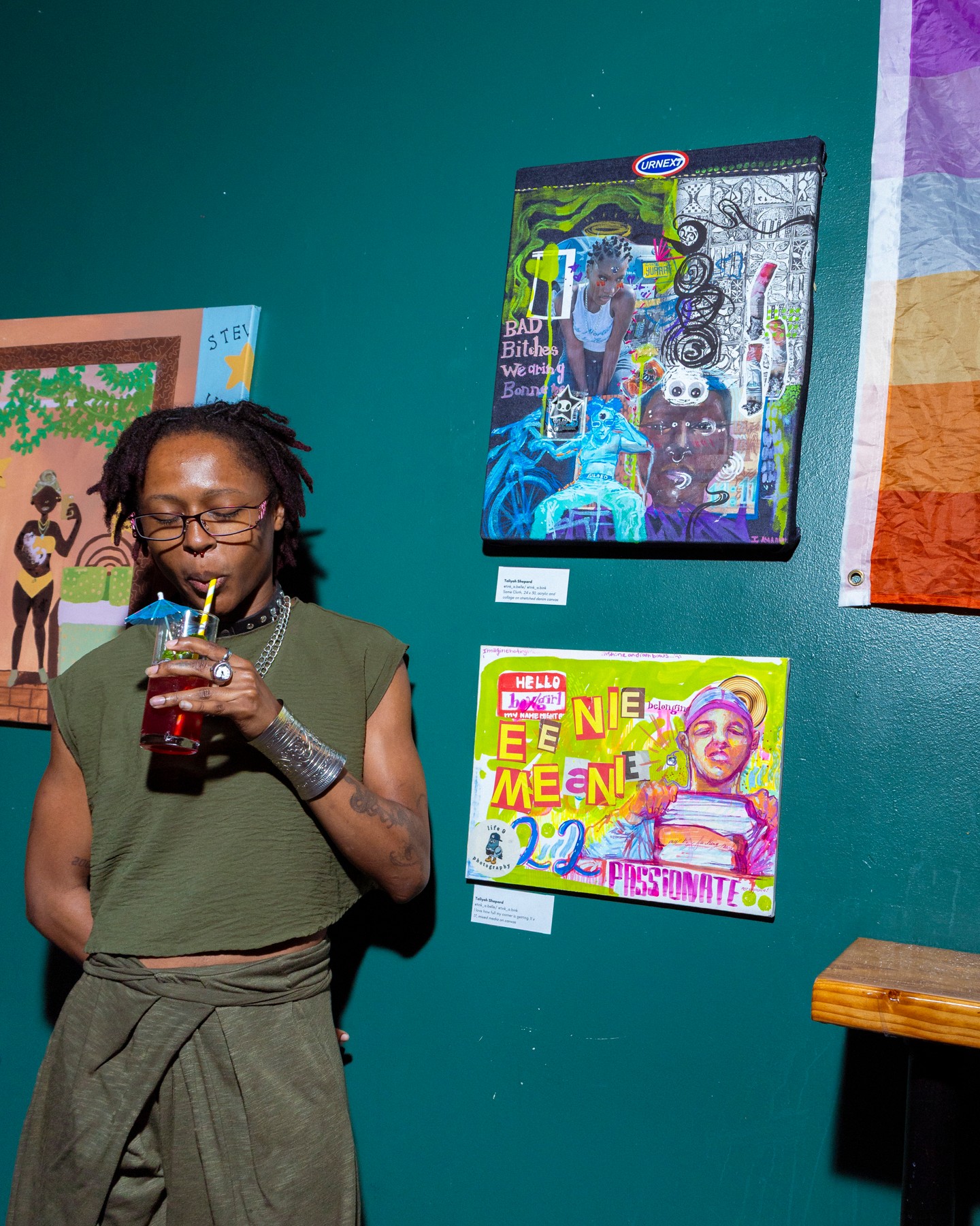 Woman with glasses and dreadlocks drinks from a cup in front of colorful artwork on a teal wall.