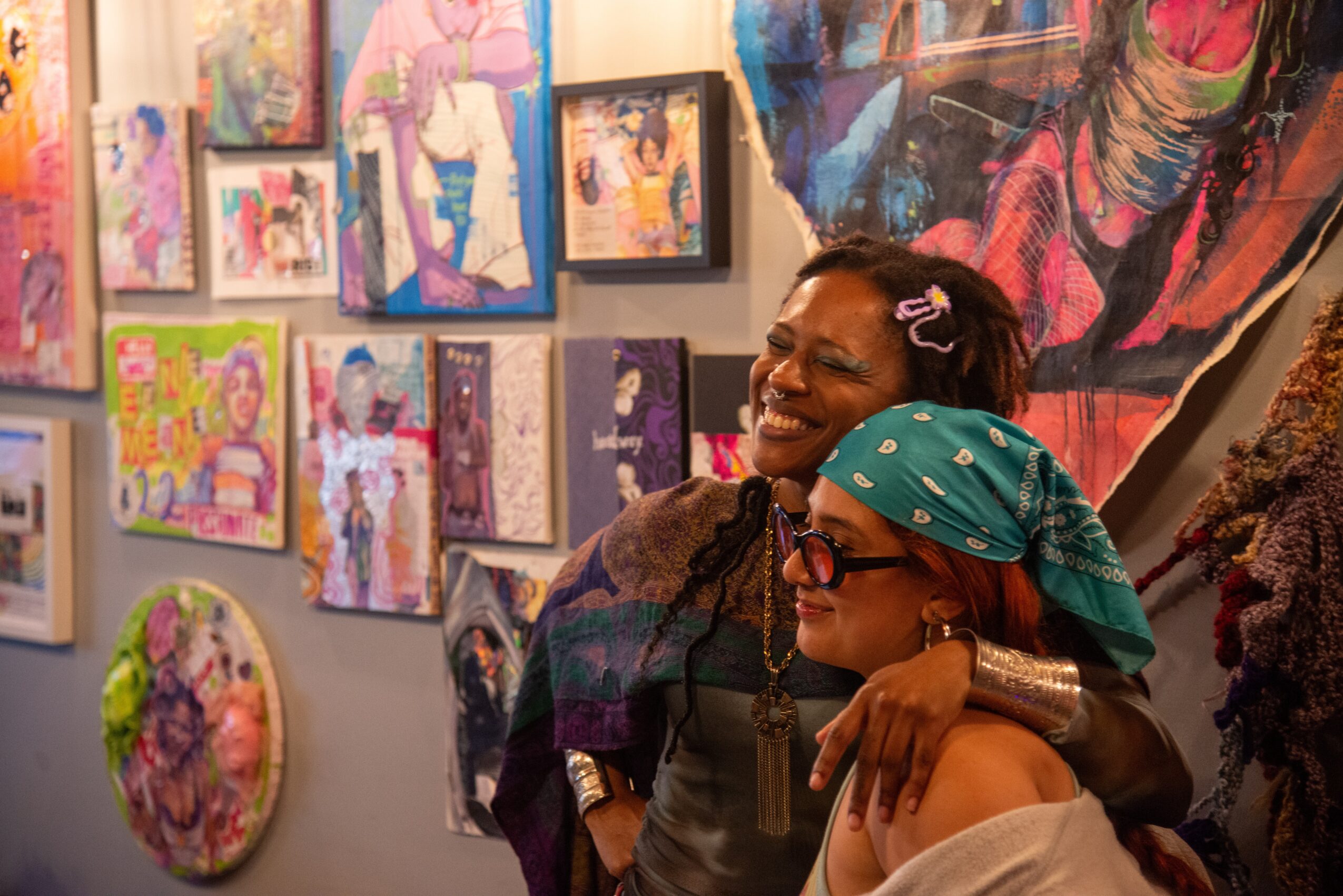 Two women embrace and smile at an art gallery with colorful paintings on the wall behind them.