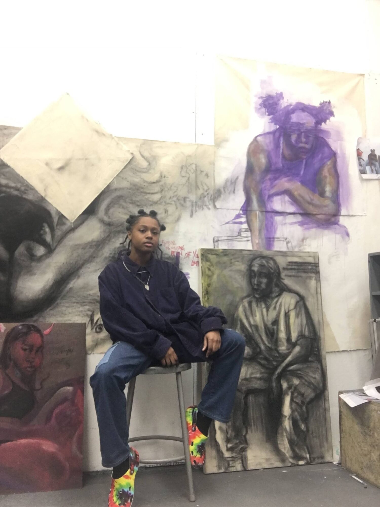 Person sitting on a chair in front of artwork, with paintings and sketches on the wall behind them.