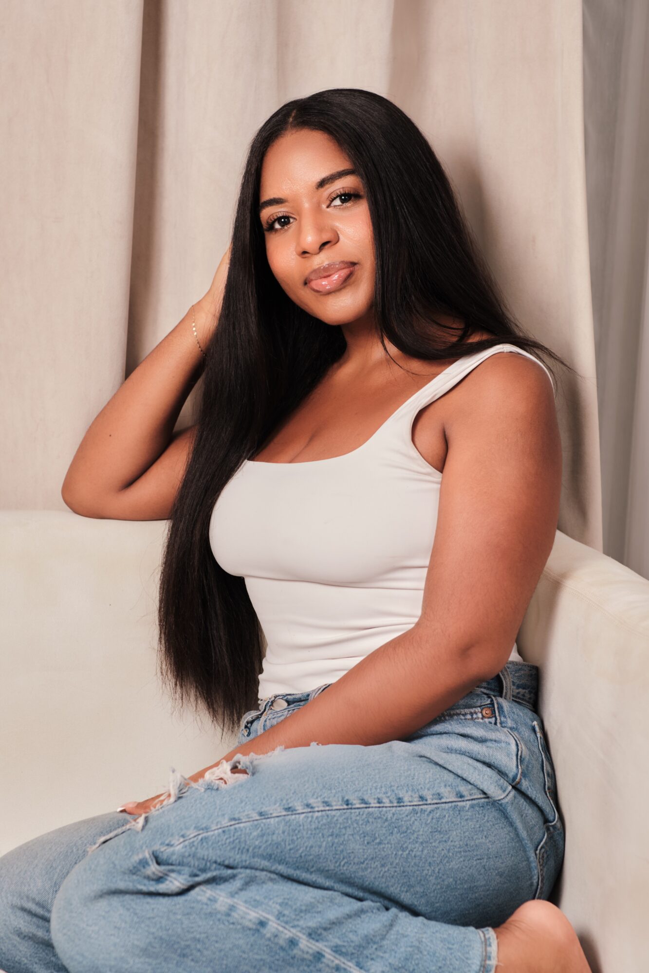 Young woman with long black hair sitting on a sofa, wearing a white tank top and ripped jeans, looking at the camera.