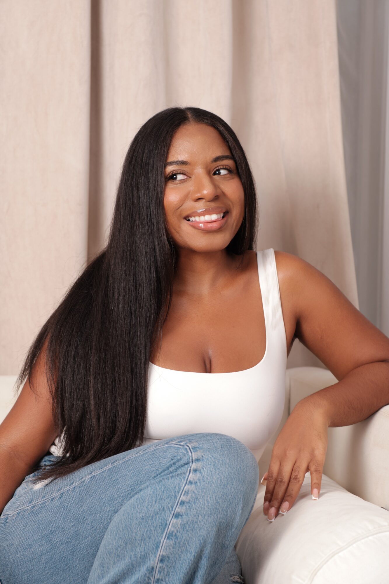 Young woman with long black hair smiling, wearing a white top, sitting on a white couch, beige curtains in background.