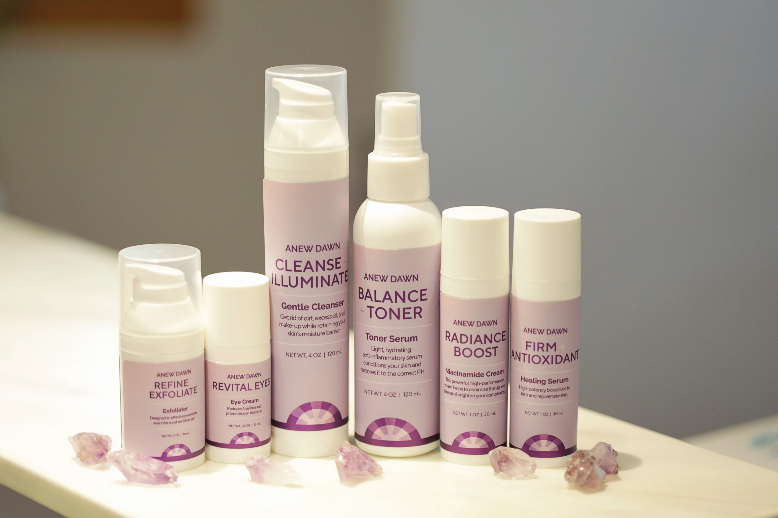 Six skincare bottles arranged on a surface with small pink crystals, against a neutral background.
