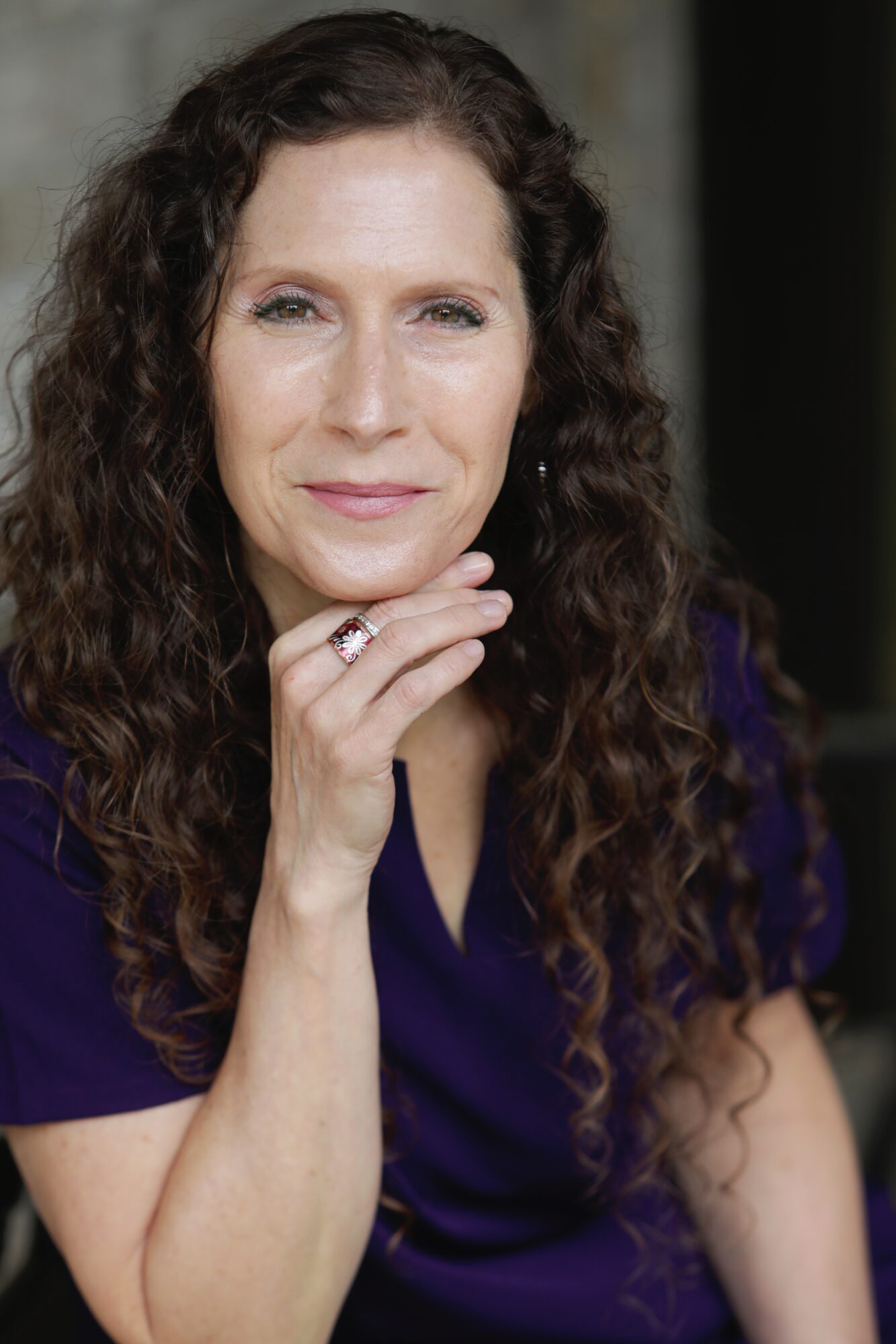 Woman with long curly hair, wearing a dark purple top, resting her chin on her hand, smiling softly.