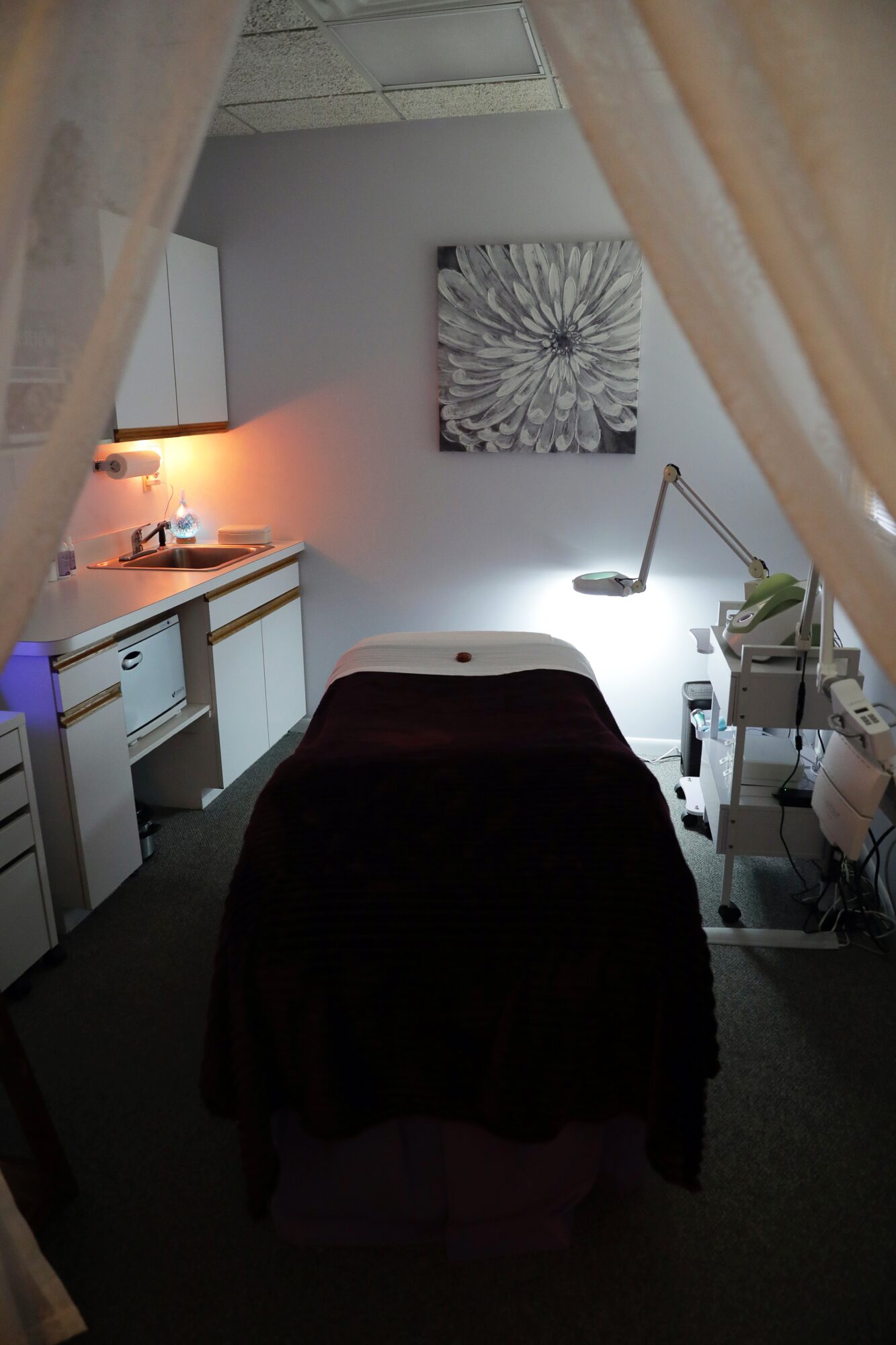 Treatment room with a bed covered in dark fabric, a desk lamp, and a floral artwork on the wall.