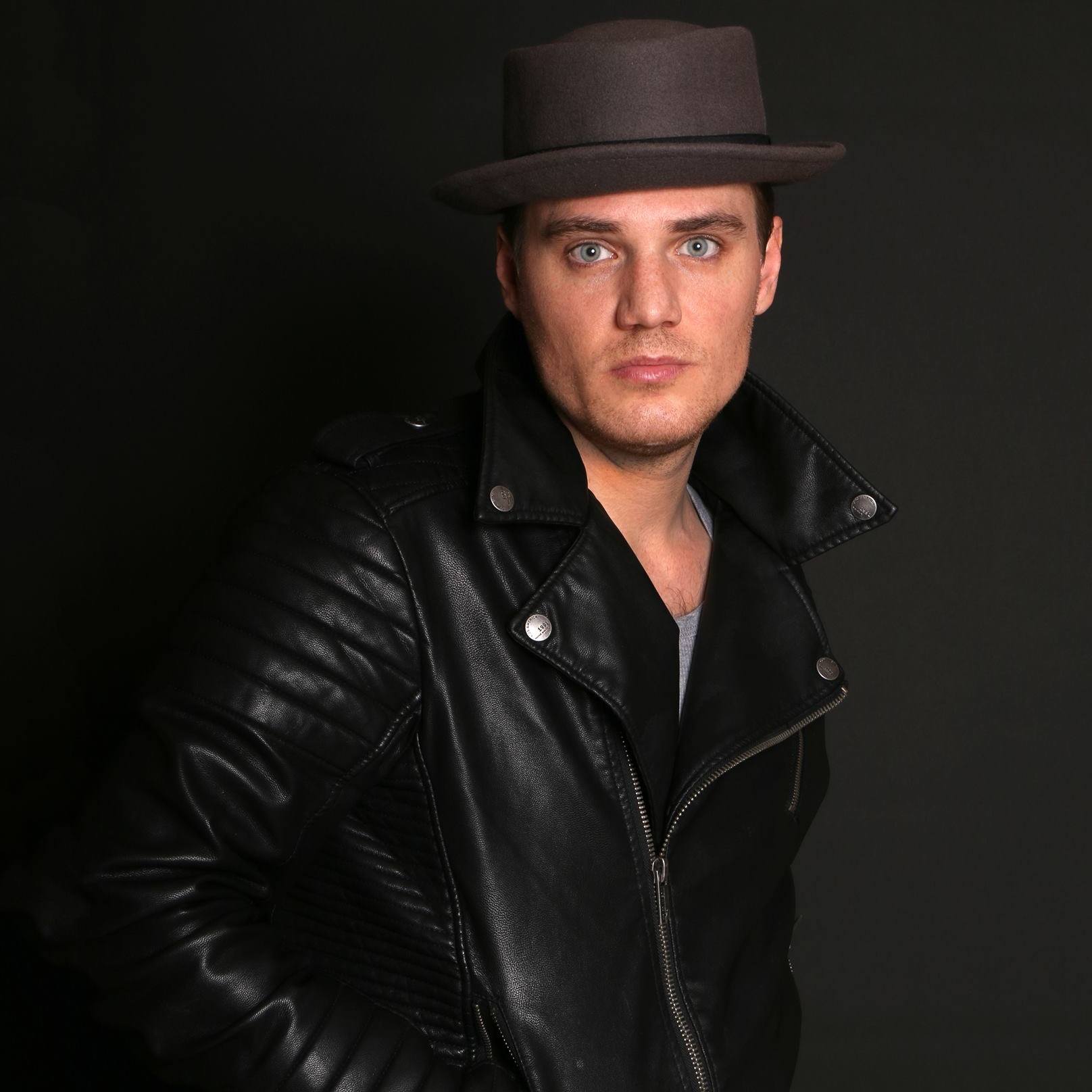 Young man wearing a black leather jacket and a wide-brimmed hat, looking directly at the camera against a dark background.