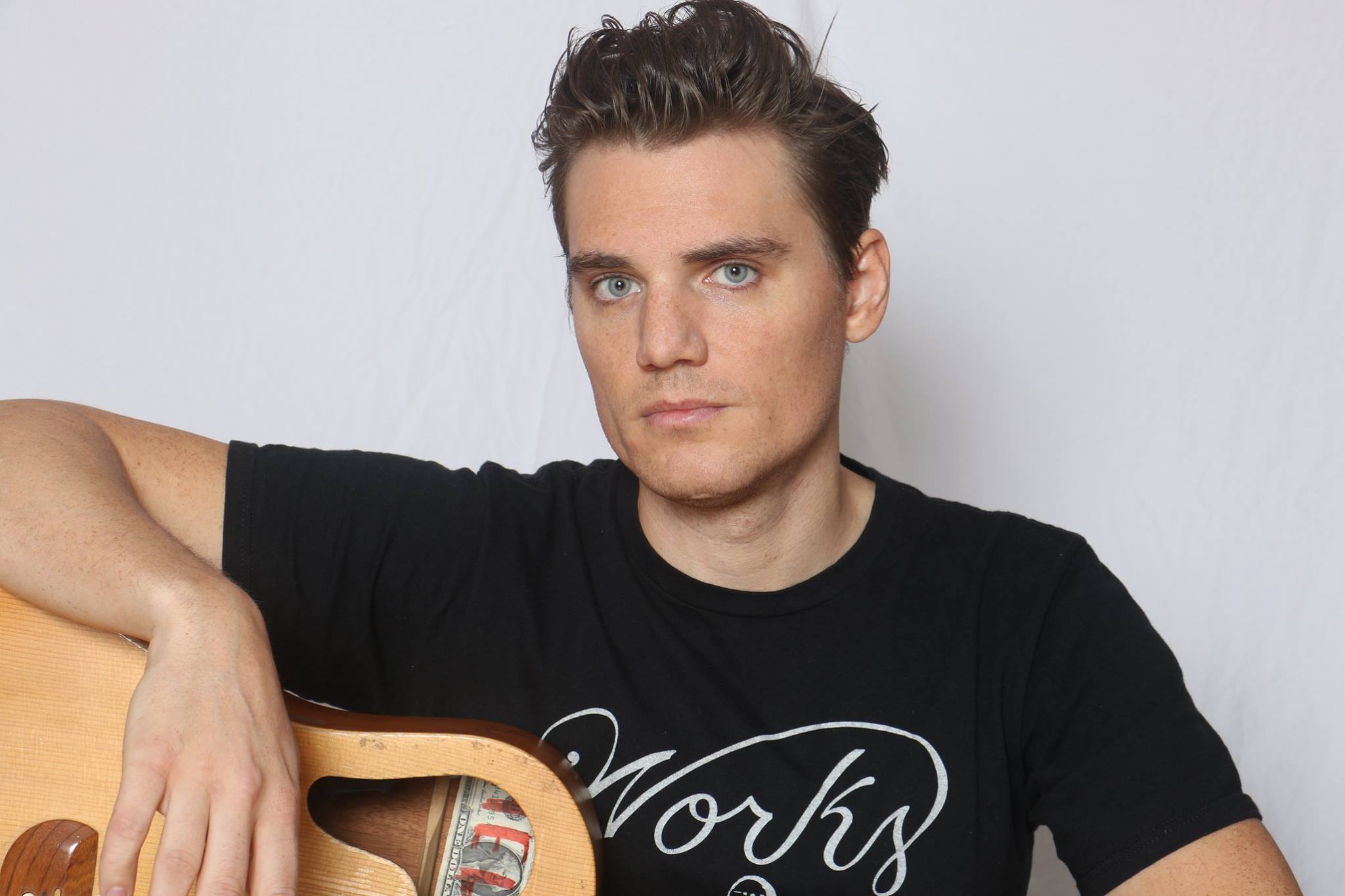 Young man with dark hair and blue eyes wearing a black T-shirt, sitting with his arm resting on a guitar, against a plain white background.