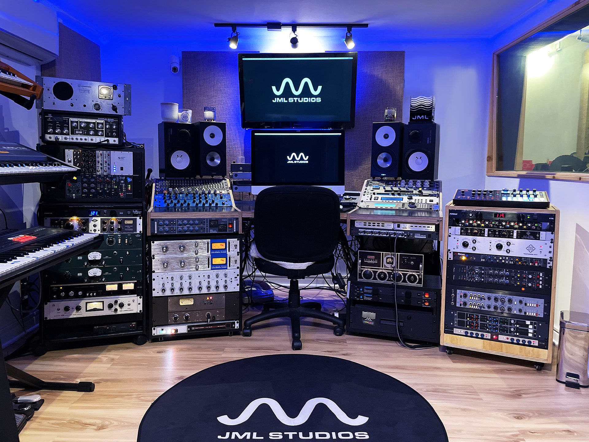 Music studio with speakers, mixing equipment, and a monitor on the wall, illuminated by blue lighting, with a black chair and a round rug.