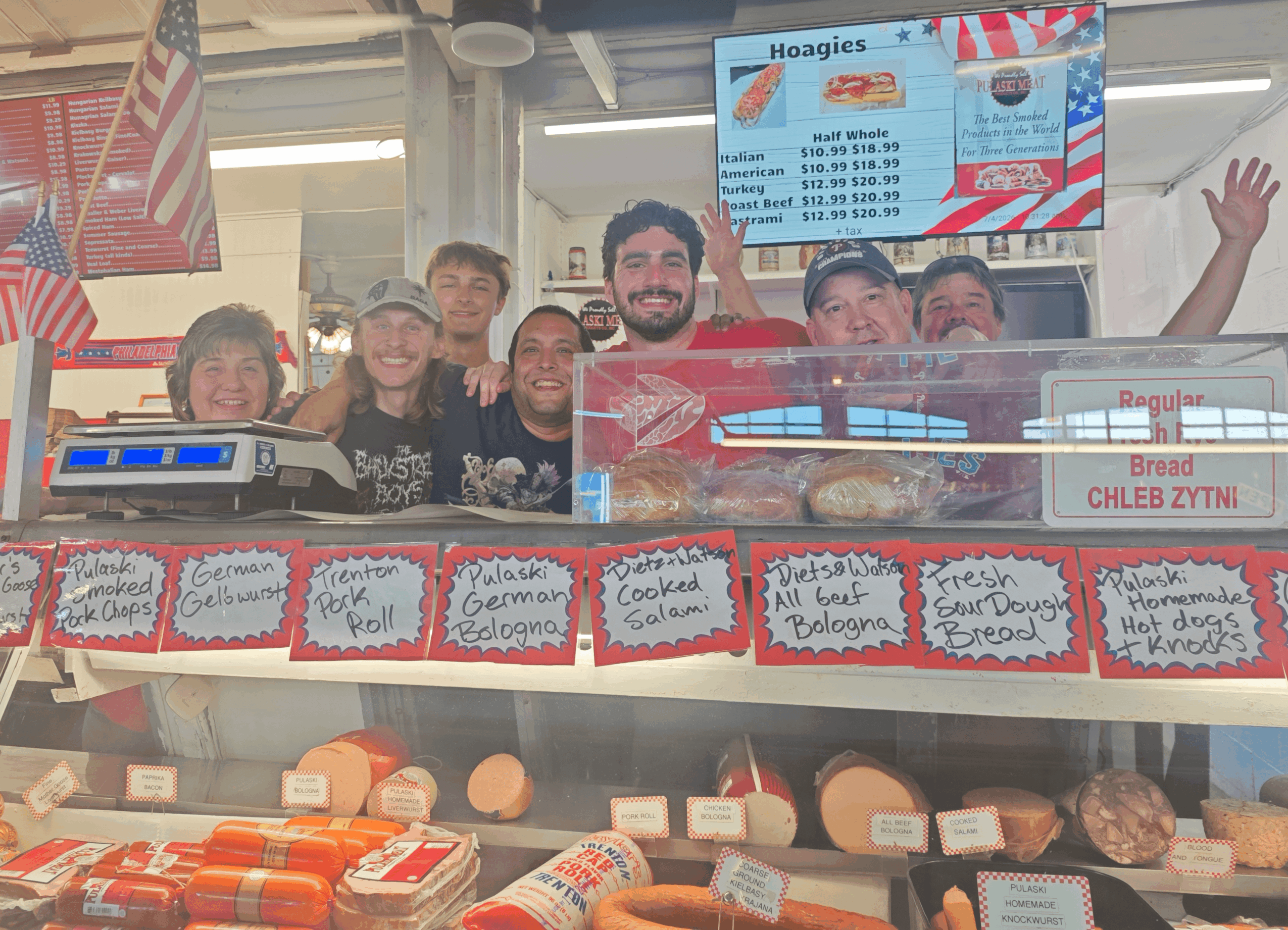 Group of people behind a deli counter with signs and a menu board, smiling and waving, in a store setting.