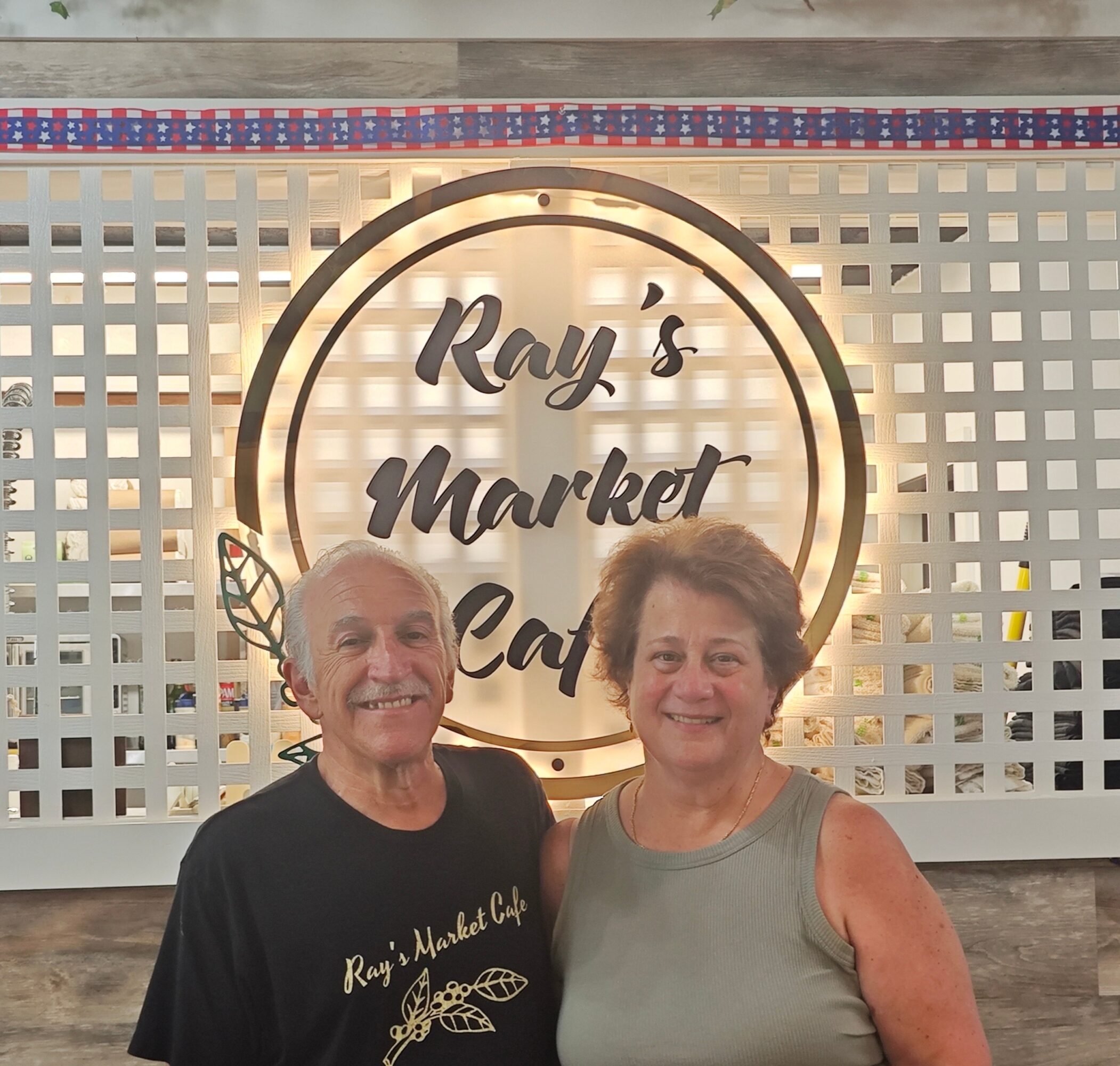 Two smiling people standing in front of a sign that reads 'Ray's Market Cafe' with a decorative background.