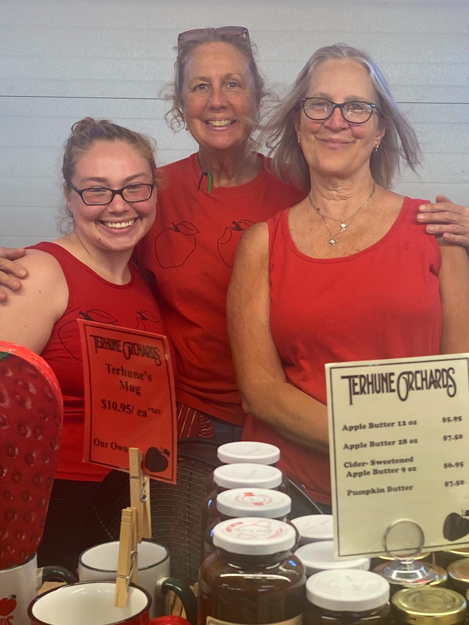 Three women in red tops smiling behind a table with jars and signs, indoors with a light-colored wall.